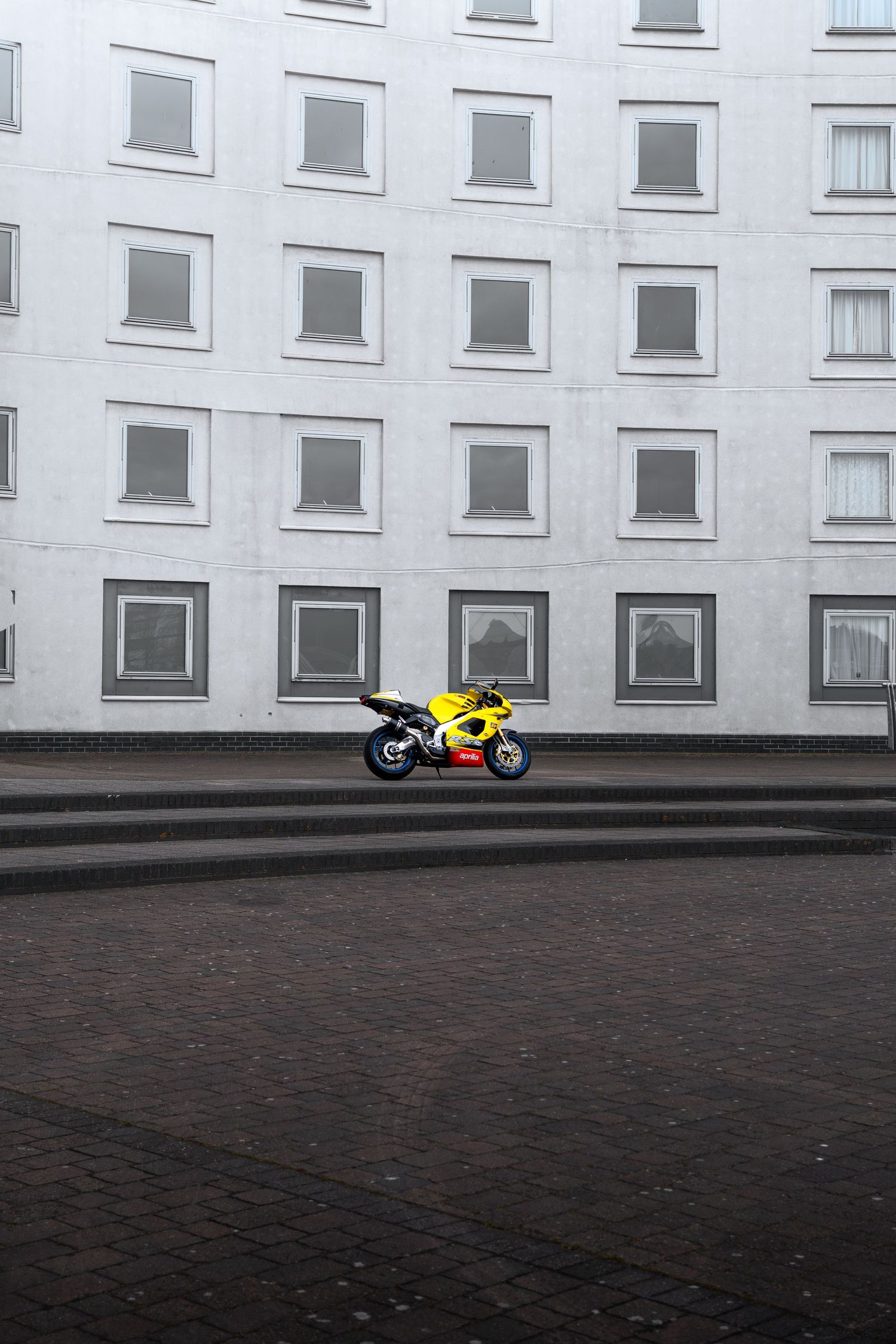 Yellow Aprilia RSV sport motorcycle parked on cobblestones against a large white building.