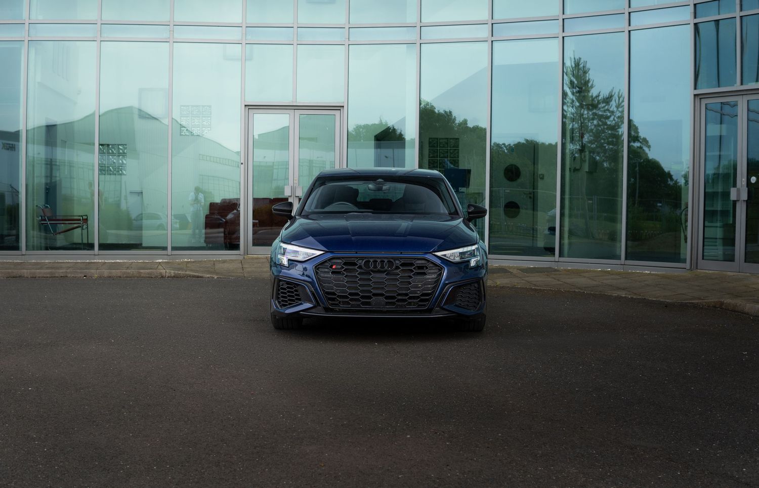 Navy blue Audi S3 sedan parked in front of a modern glass building at dusk.