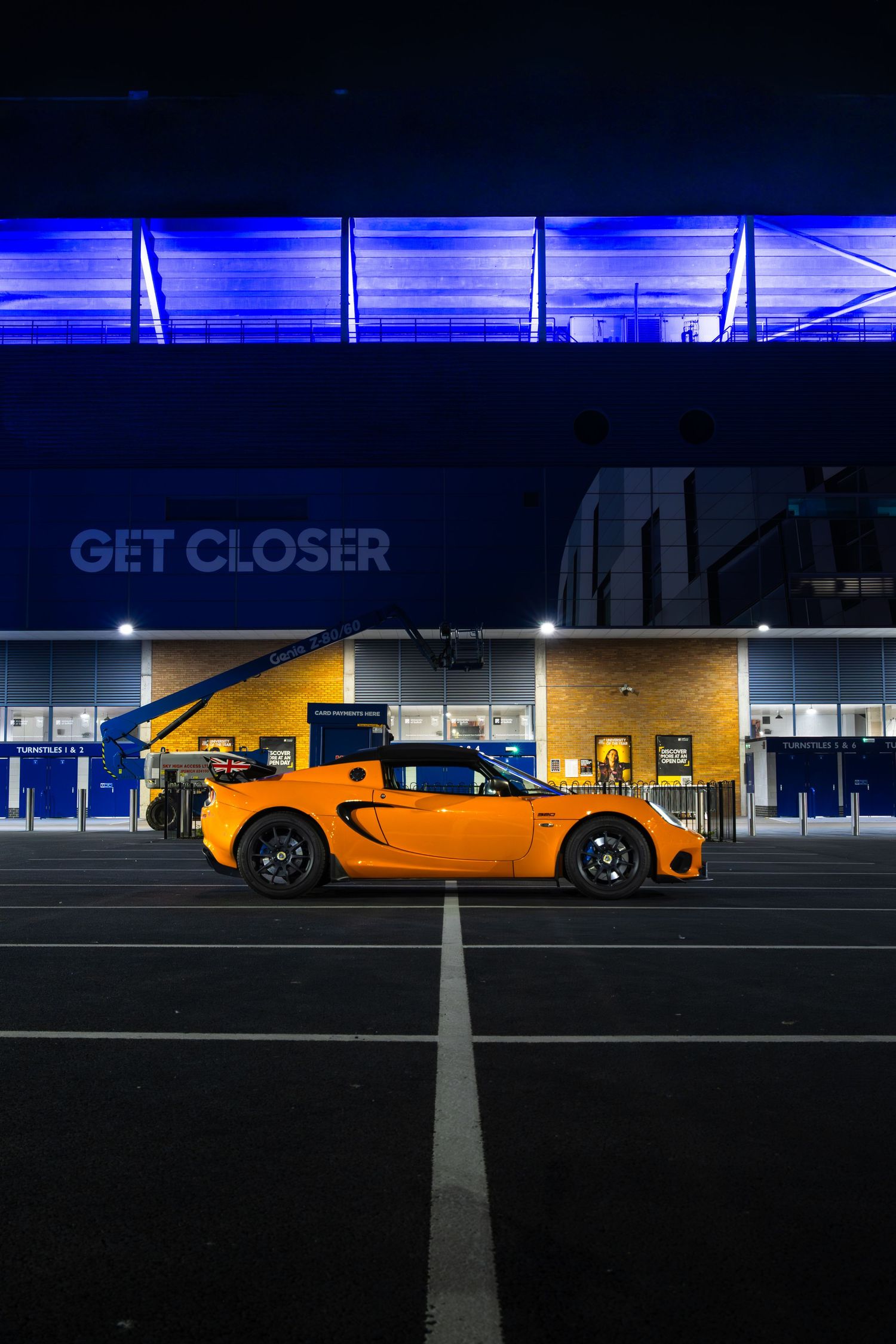 Low-angle side view of orange Lotus Elise in empty car park outside blue-lit stadium GET CLOSER entrance at night.