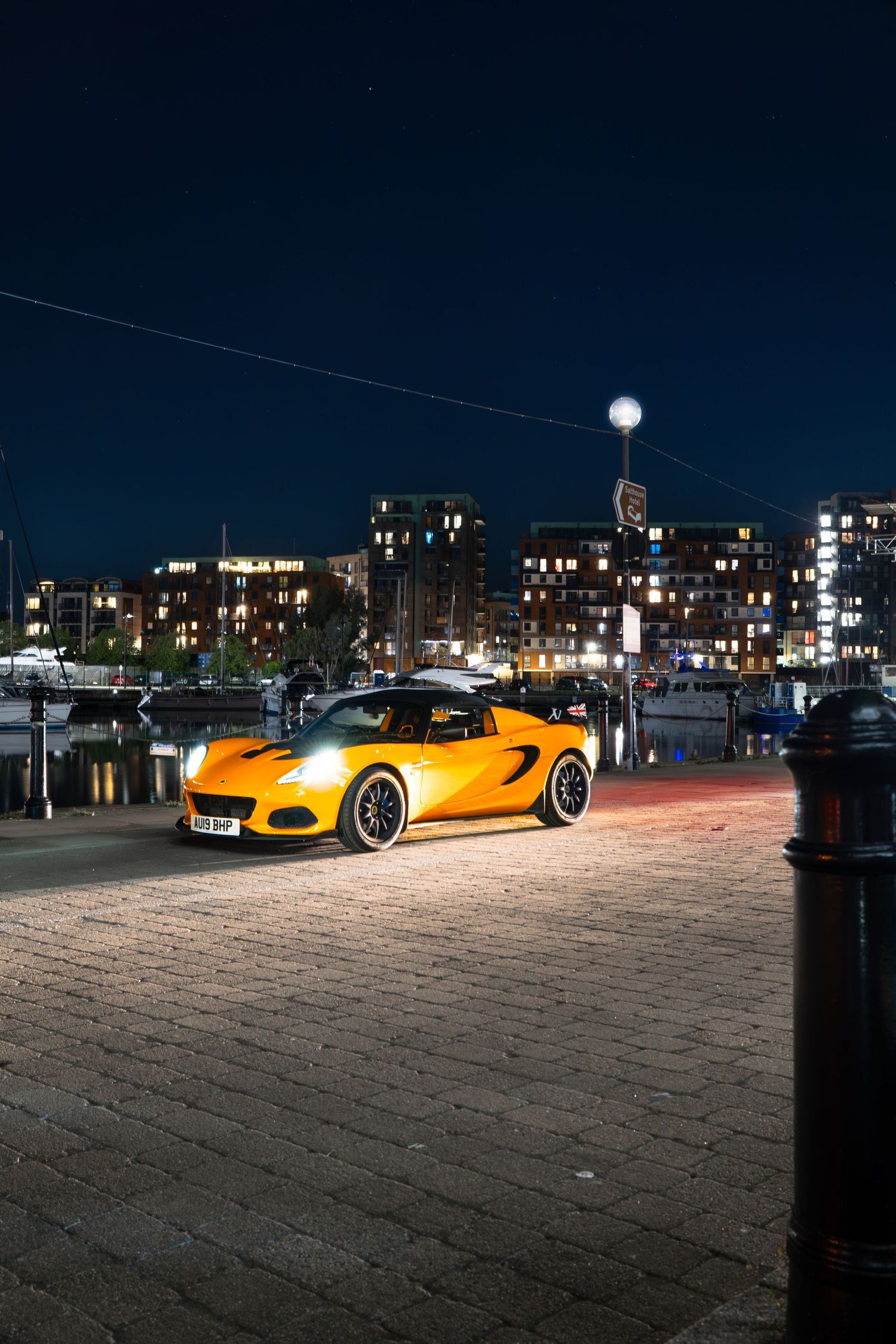 Yellow Lotus Elise sports car on cobblestone quayside at night with marina and lit buildings behind.