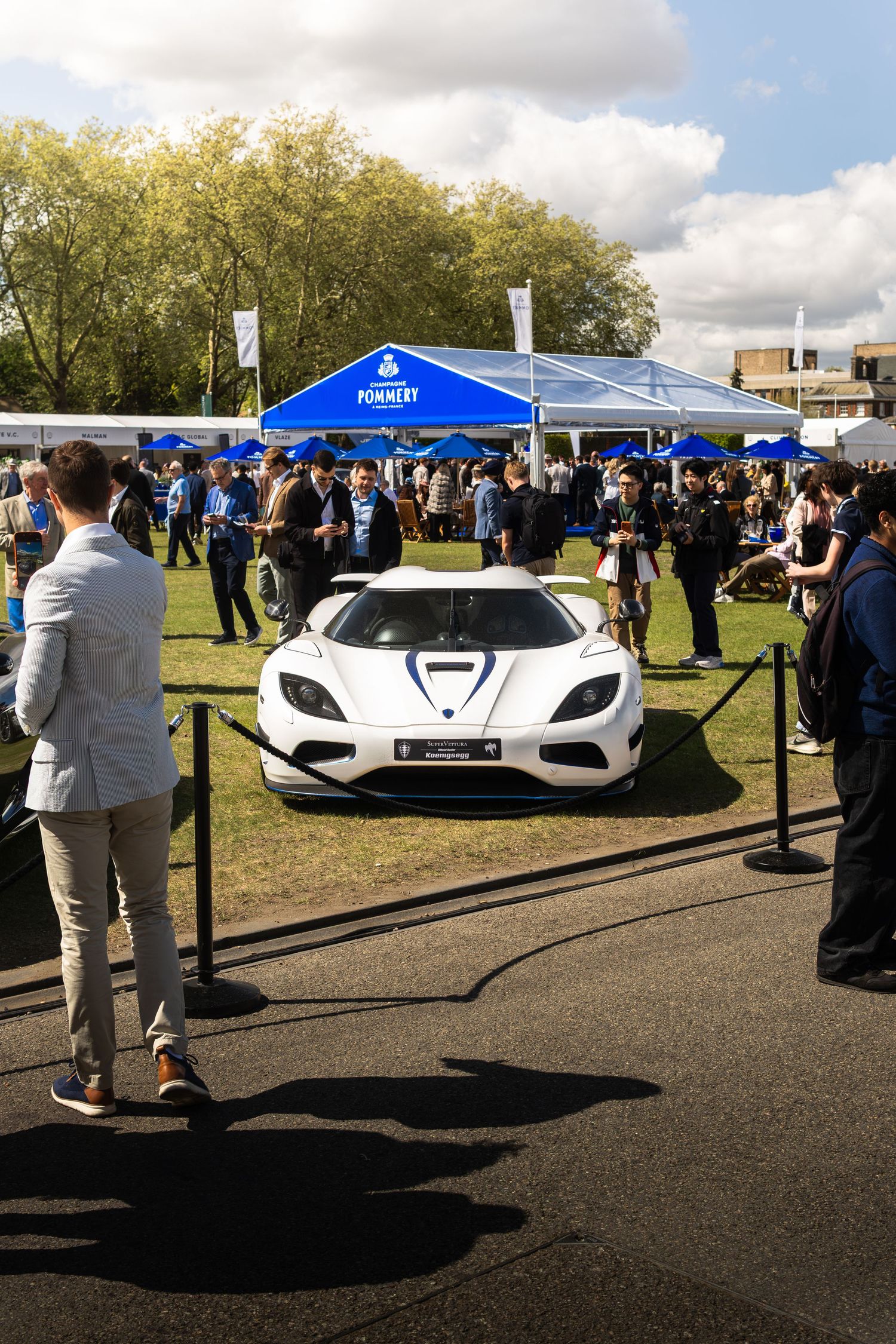 White Koenigsegg supercar on display at an outdoor luxury car show with Pommery sponsor tent.