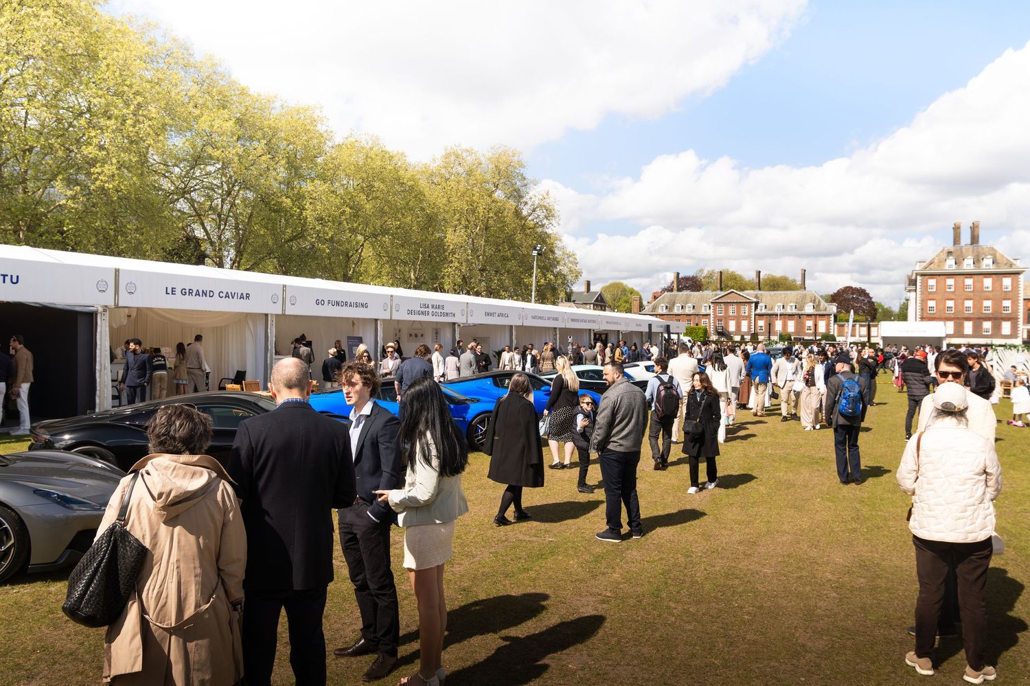 Large crowd of attendees browsing luxury cars at an outdoor automotive exhibition on a sunny day.