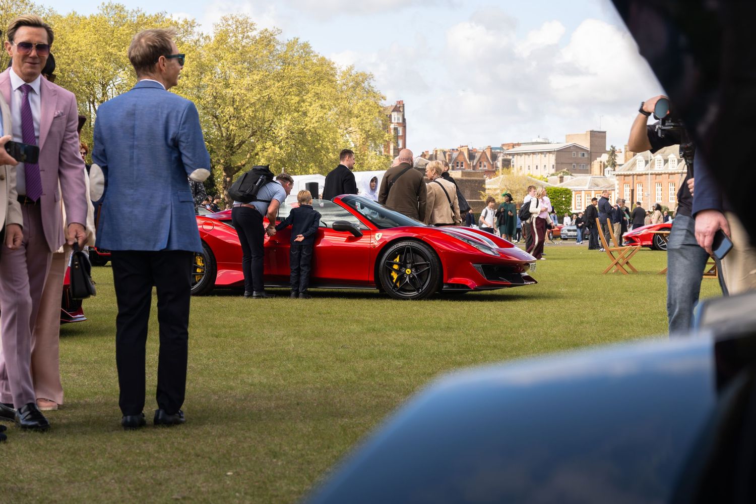 Elegantly dressed attendees admiring a red Ferrari 488 Pista at an outdoor car show on grass.