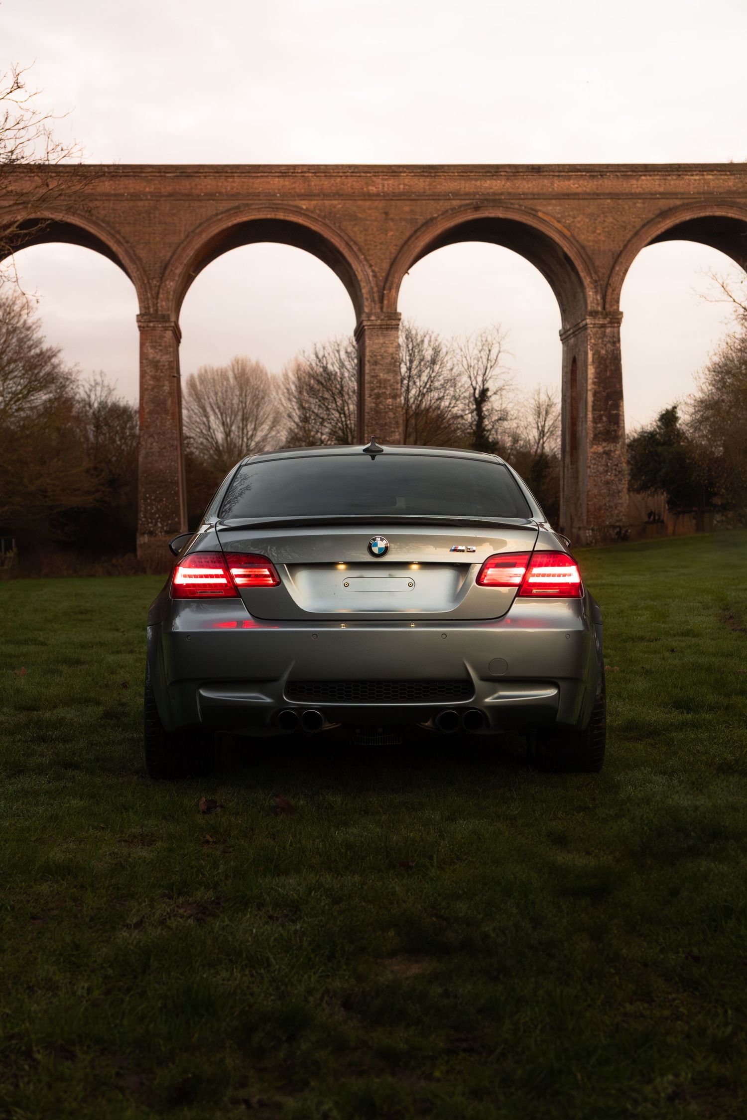 Silver BMW M3 coupe parked on green grass with a large brick arched viaduct behind it.
