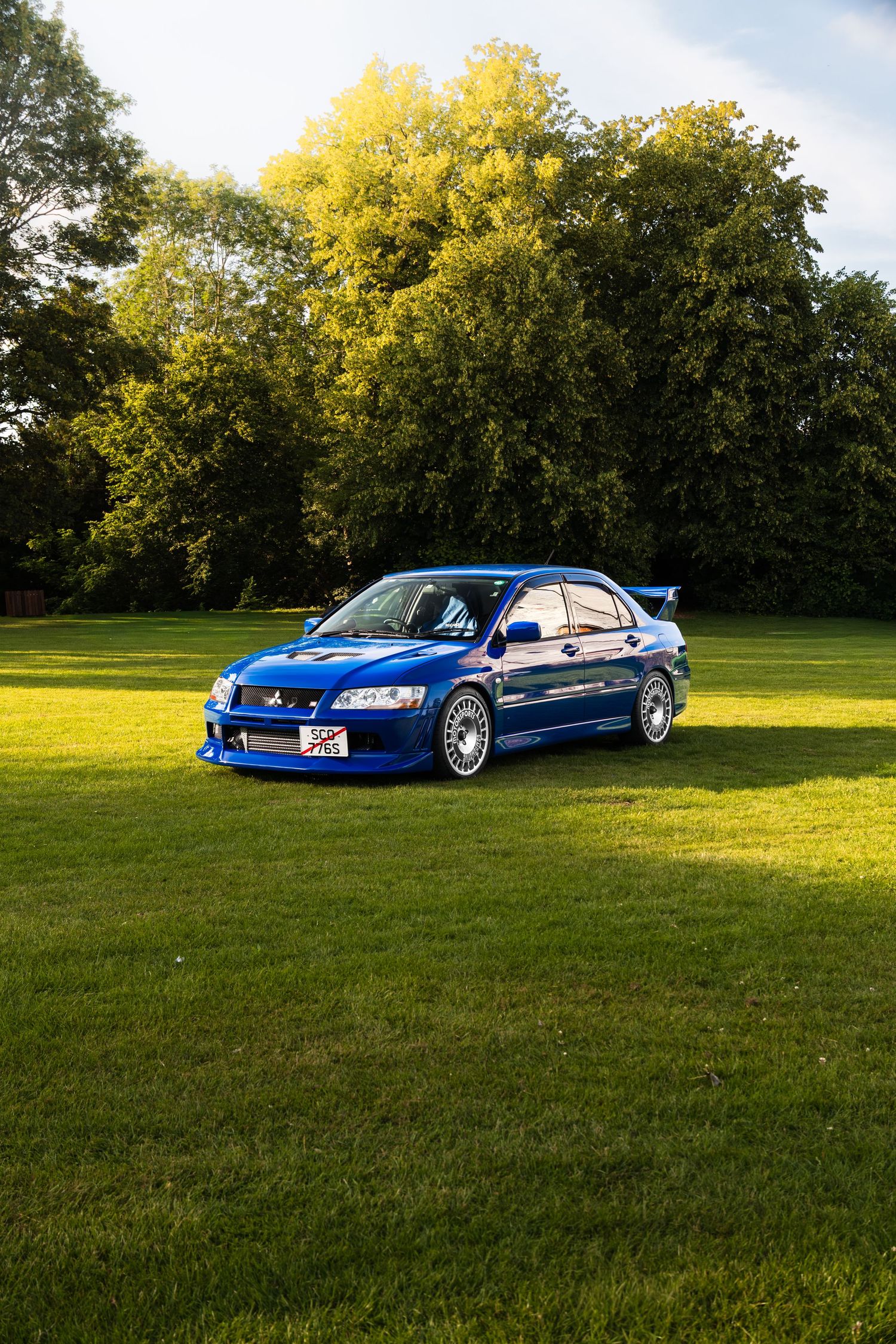 Blue Mitsubishi Lancer Evolution parked on green grass with sunlit trees in the background.
