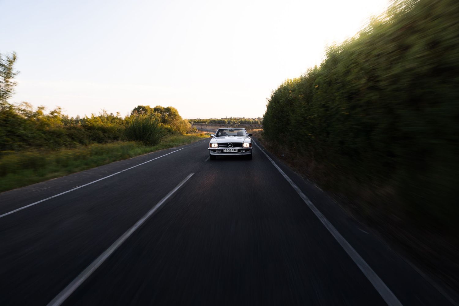White car driving on a two-lane road surrounded by lush green trees at sunset.