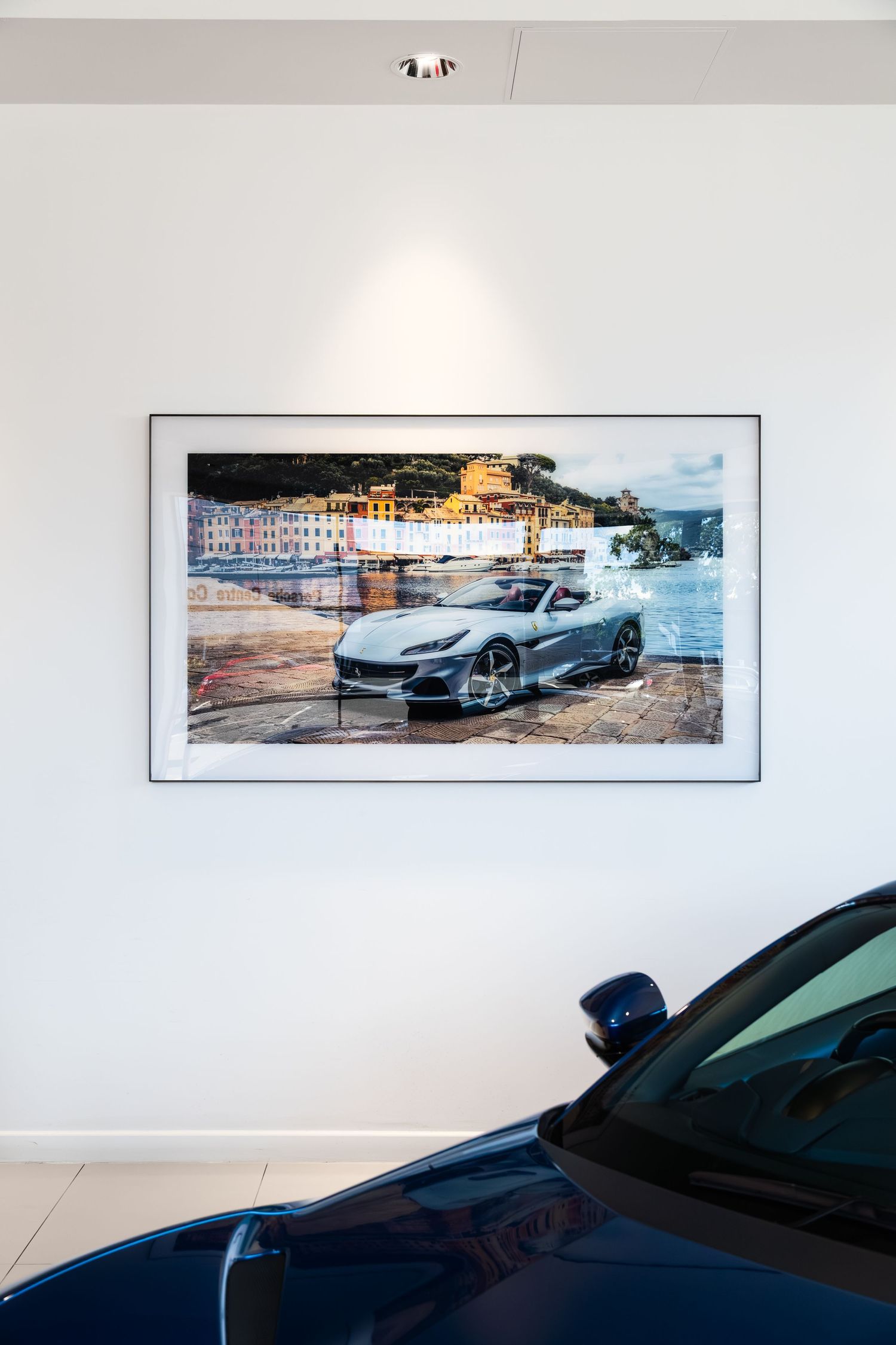 A framed photo of a silver Ferrari sports car in a coastal town hangs on a white wall above a blue car in a showroom.