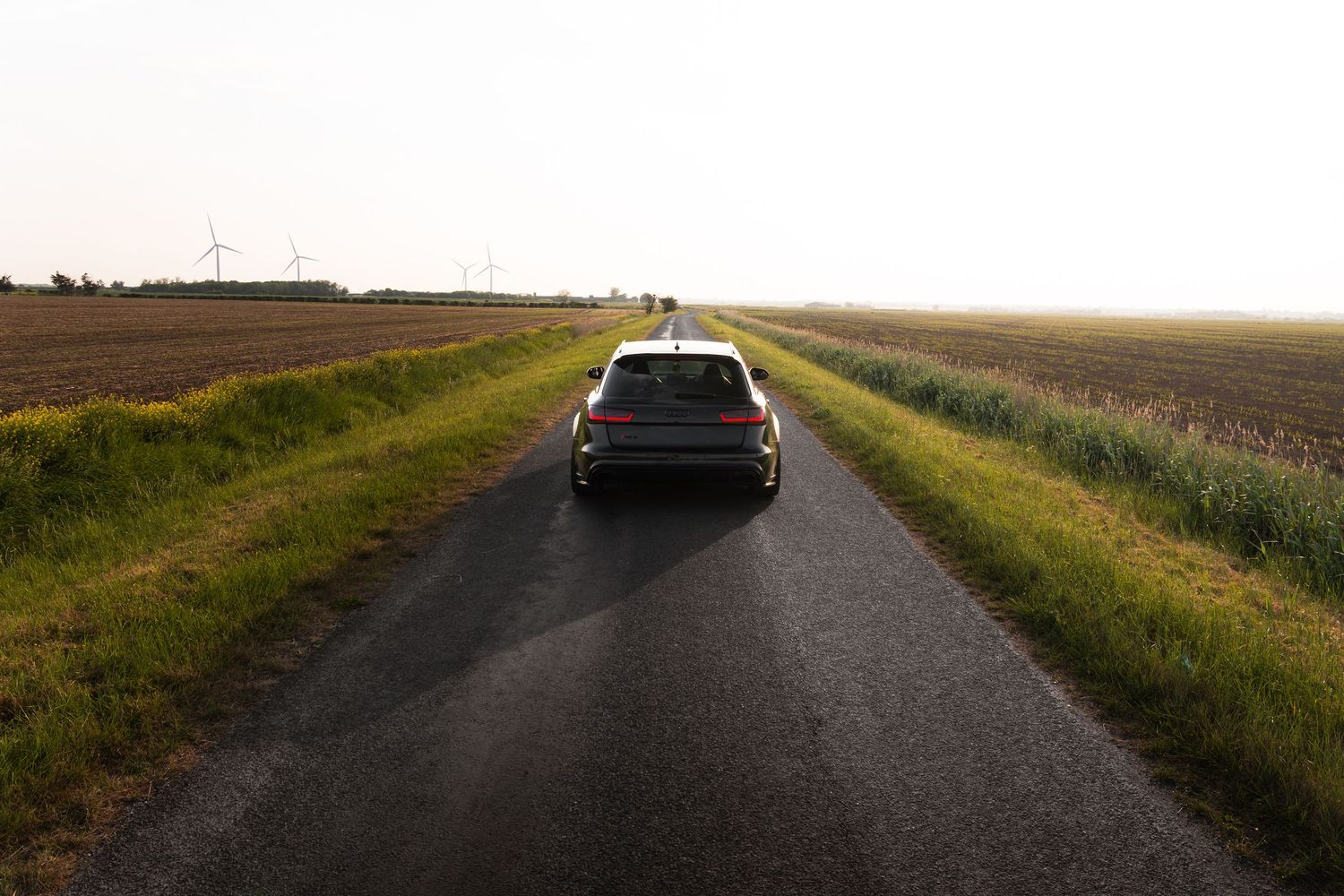 Rear view of a dark grey Audi RS6 driving on a narrow rural road flanked by green fields at sunset.