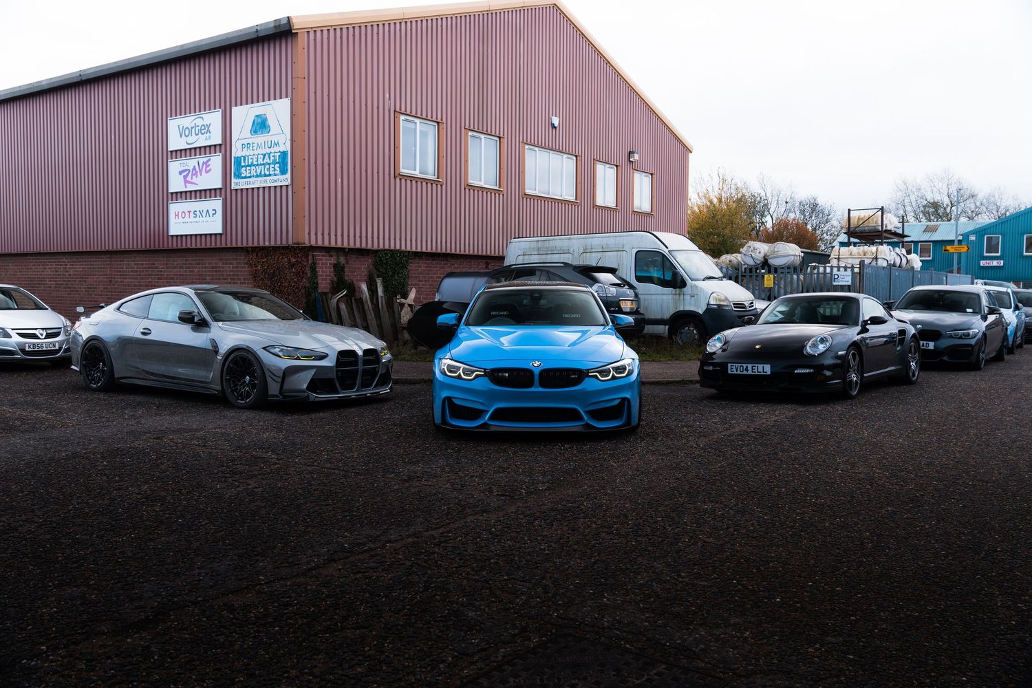 Multiple performance cars including a blue BMW M4 parked outside an industrial building.