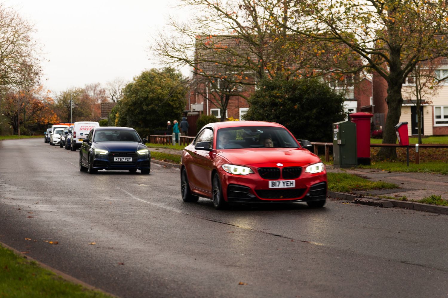 Red BMW M240i coupe leading traffic on wet suburban residential street.
