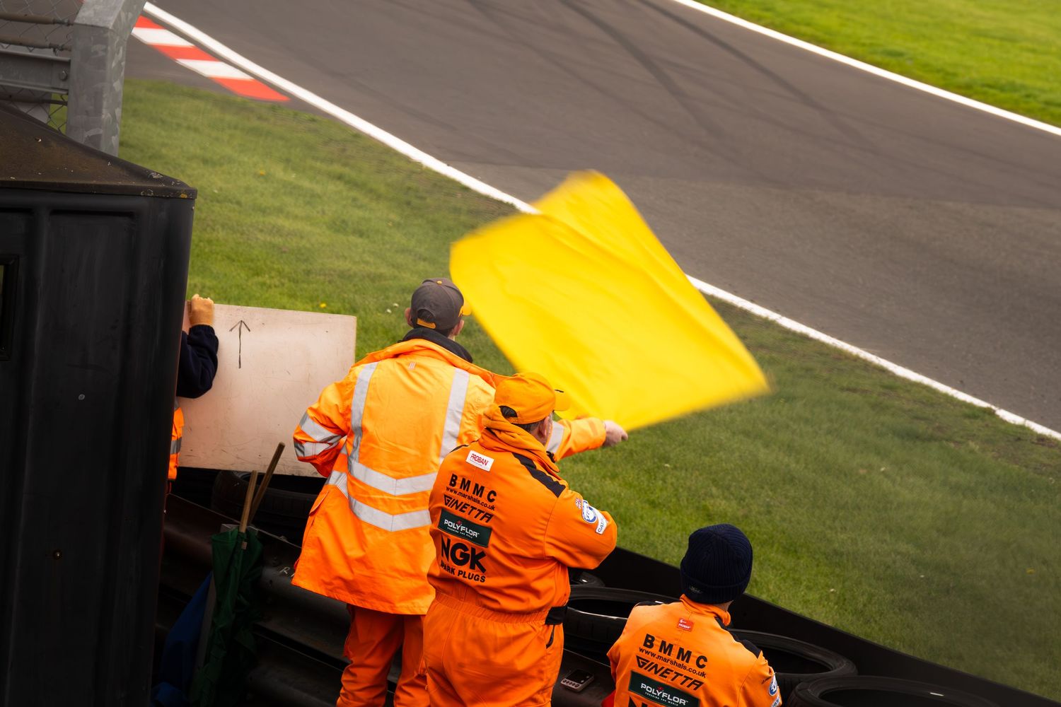 Orange-clad marshals wave a yellow caution flag trackside at a motorsport circuit.