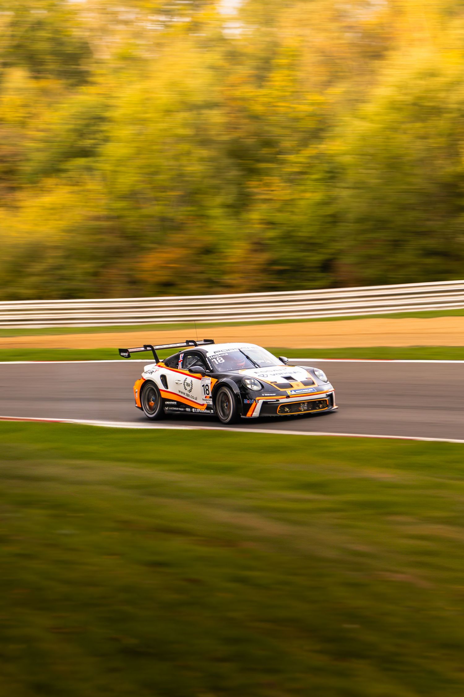 Orange and white Porsche 911 GT3 Cup race car speeding past vibrant autumn foliage on circuit.