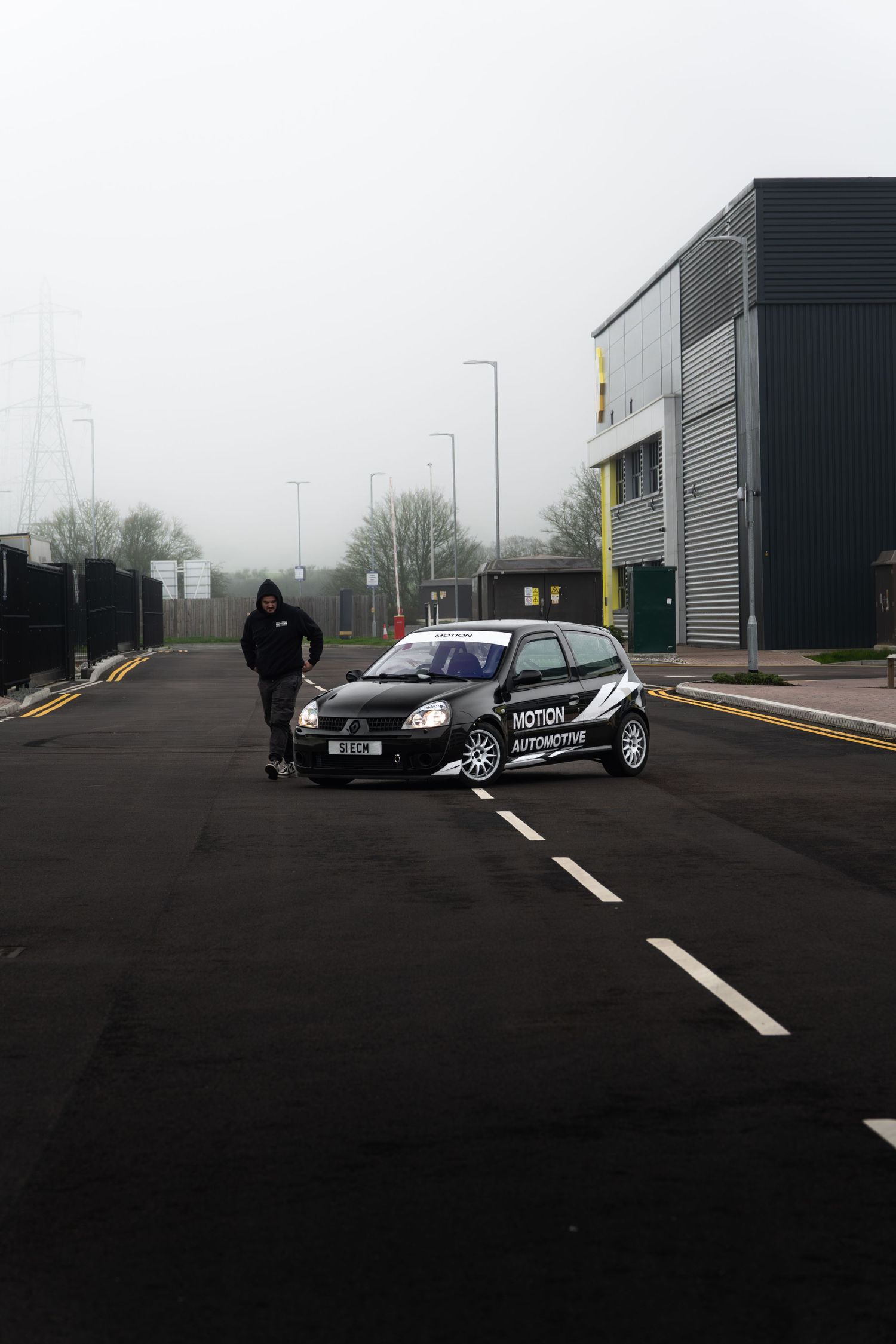 A black Motion Automotive branded Renault Clio parked on a misty industrial road with a person standing nearby.