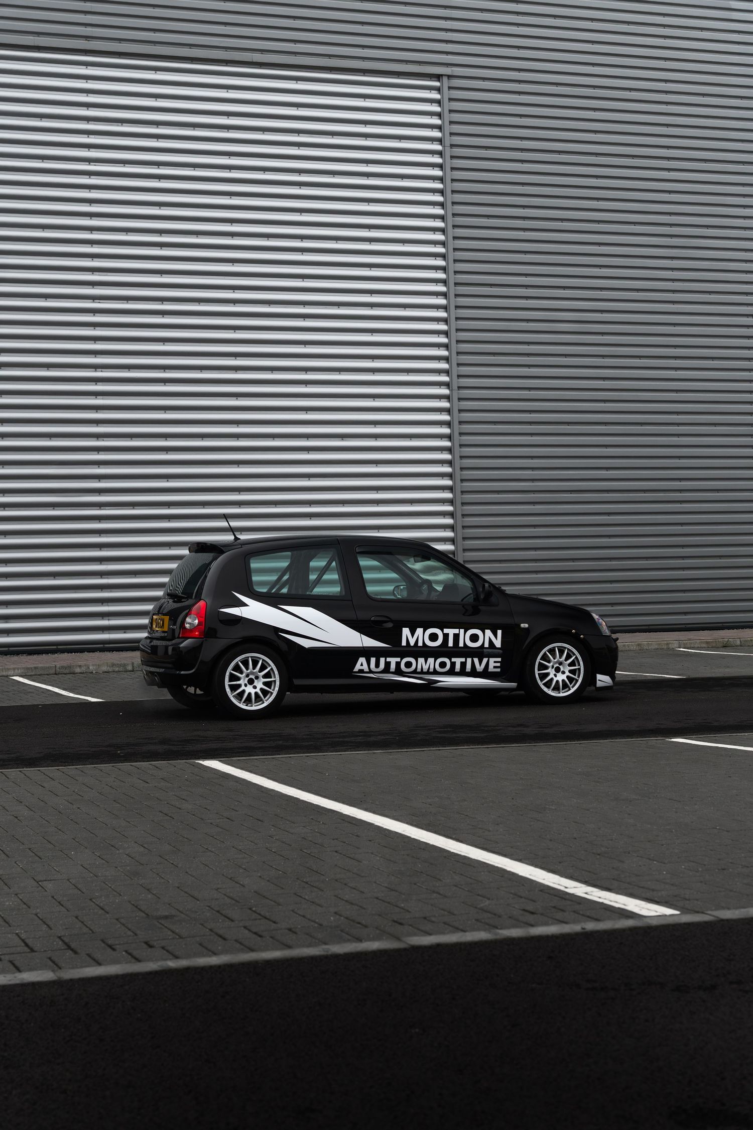 Black Motion Automotive liveried Renault Clio photographed from distance against corrugated wall.