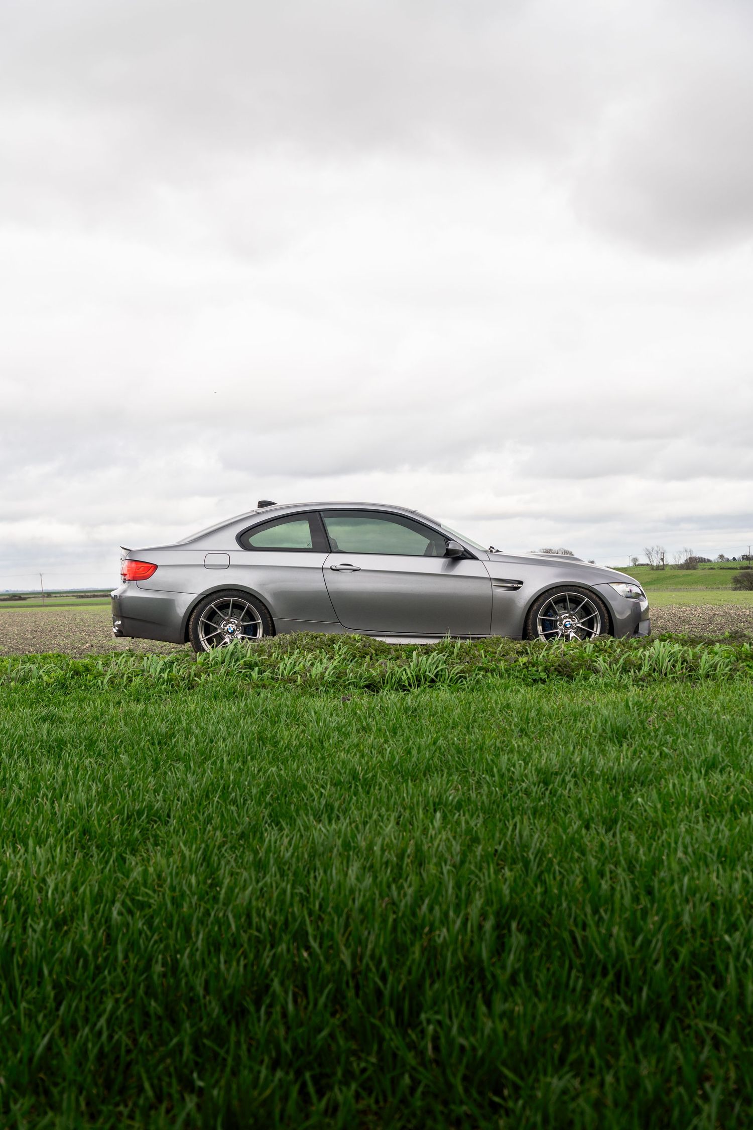 Silver BMW M3 E92 coupe parked beside a lush green field under a cloudy sky.