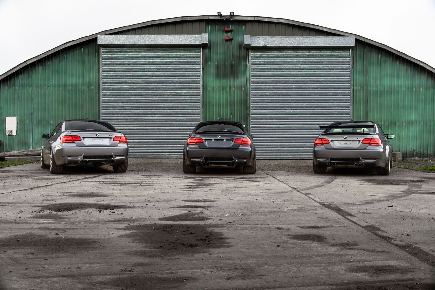 Three BMW M3 sedans parked in a row facing away from camera in front of a green industrial warehouse.
