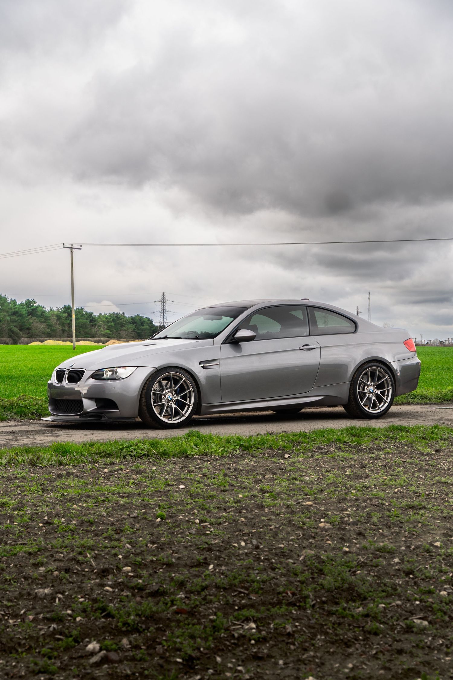 Silver BMW M3 E92 coupe parked on a gravel road beside a green field under stormy skies.