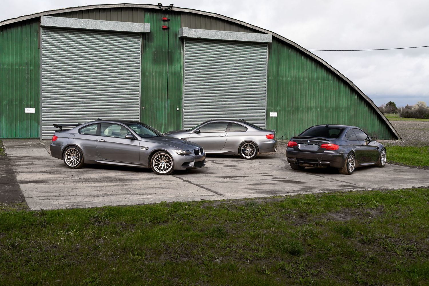 Three gray BMW M3 coupes parked on concrete in front of a large green arched hangar.
