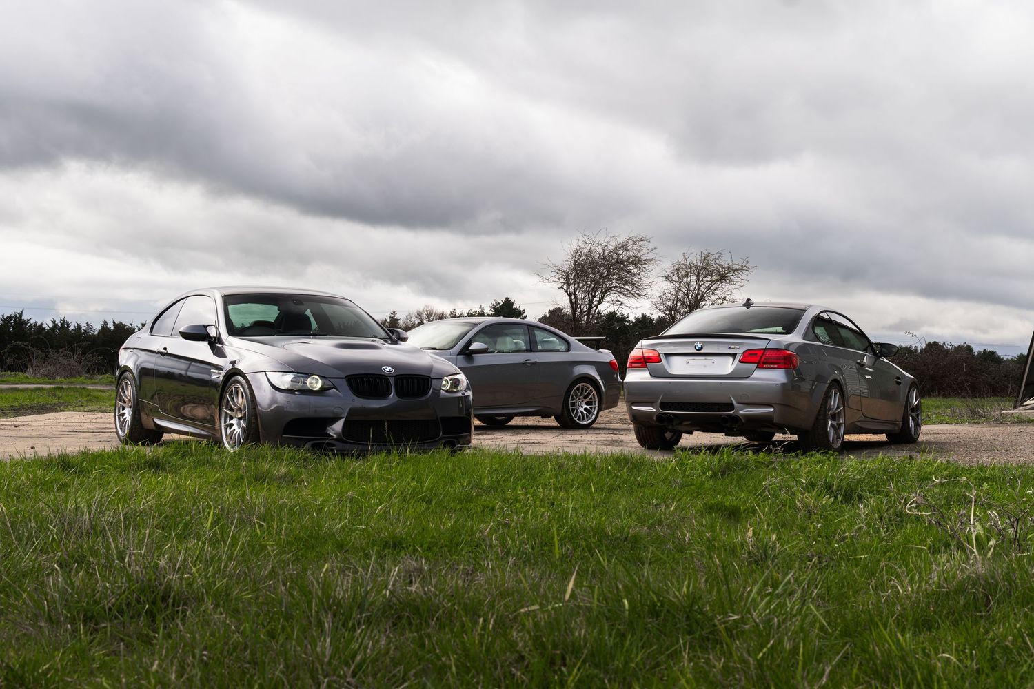Three gray BMW M3 coupes parked on grass and pavement under a cloudy sky outdoors.