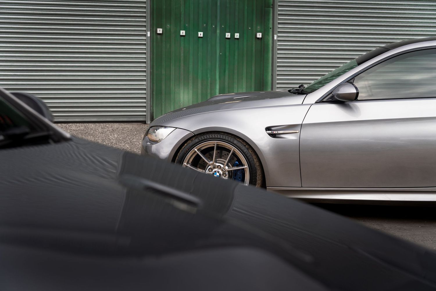 Silver BMW M3 coupe with bronze alloy wheels parked beside green garage doors.