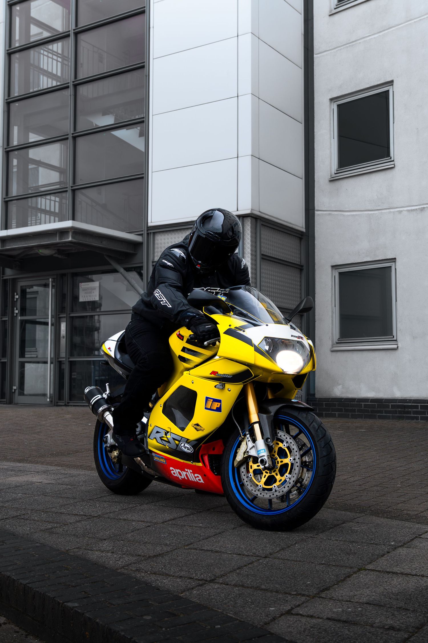 Rider on yellow Aprilia RSV sportbike with blue wheels parked near modern building.