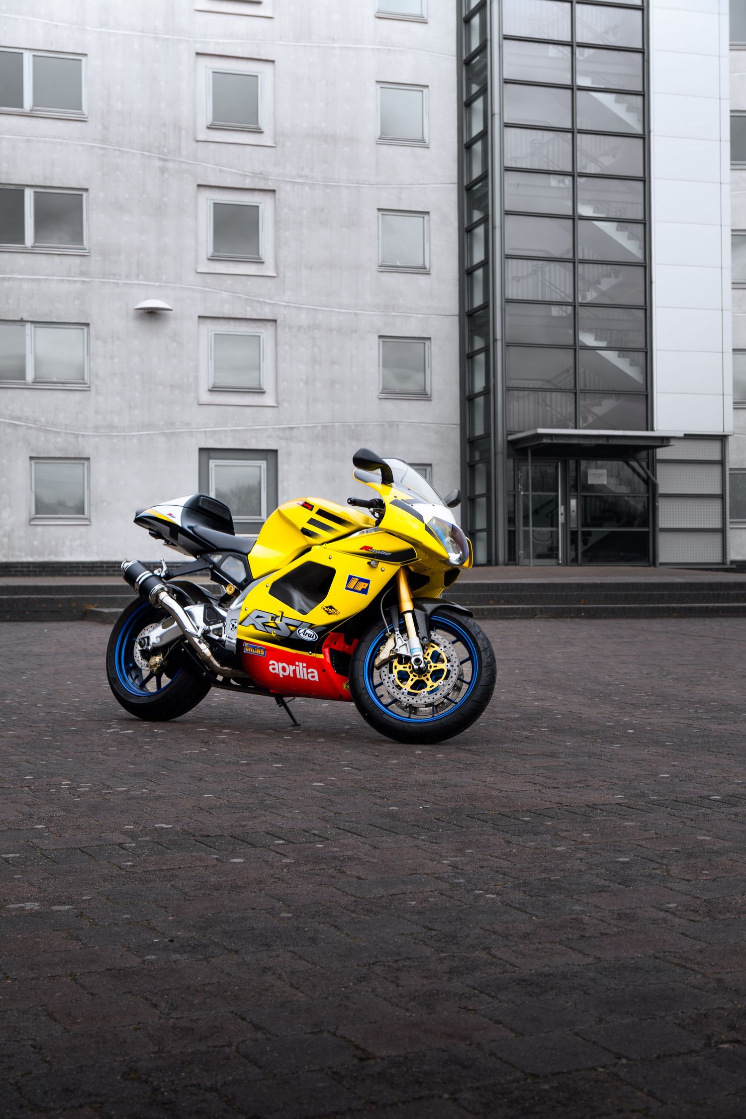 Yellow Aprilia RSV sportbike parked on cobblestones with a tall modern glass and concrete building behind.