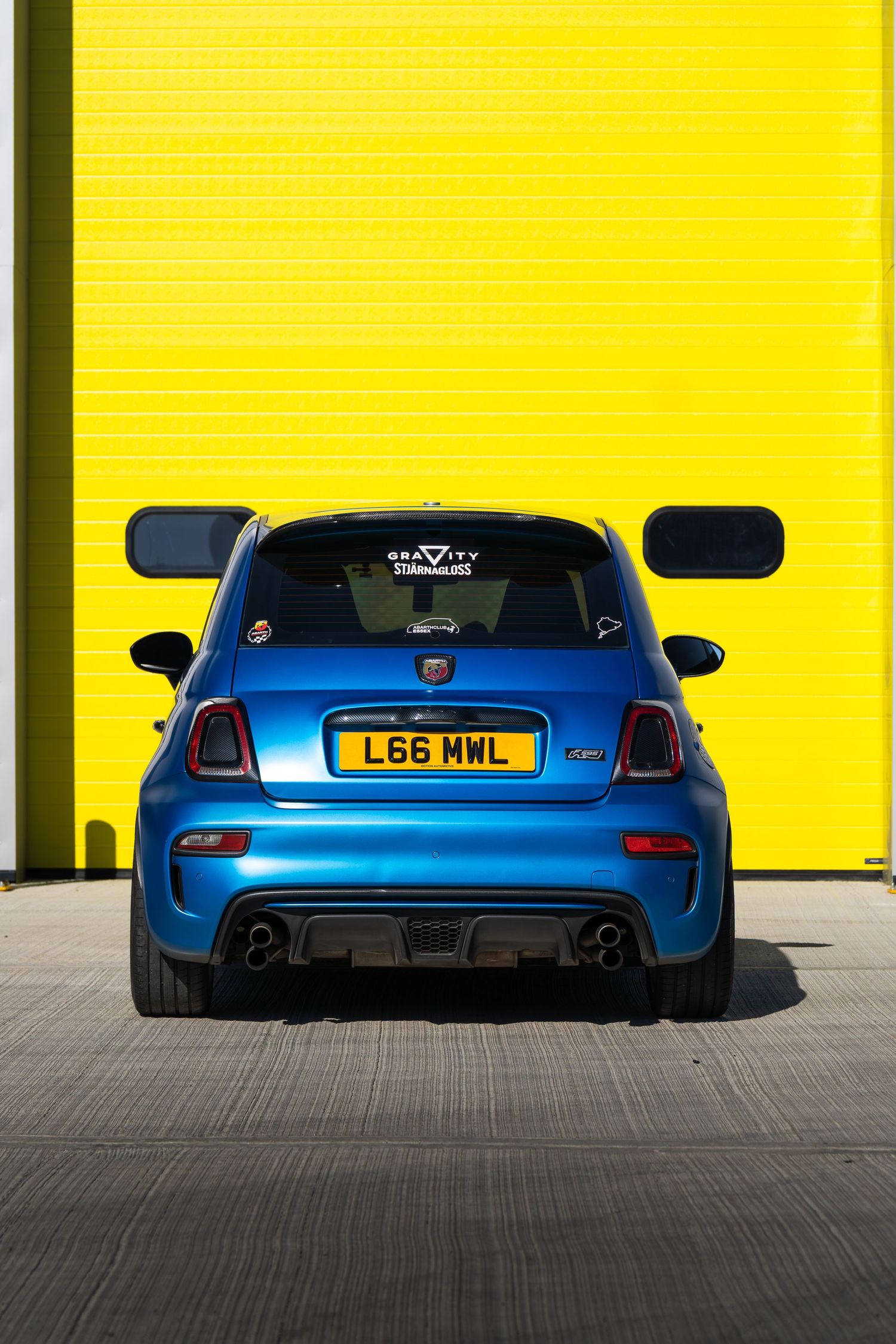 Rear view of a blue Abarth 595 with Gravity Stjarnagloss decal against a vivid yellow door.
