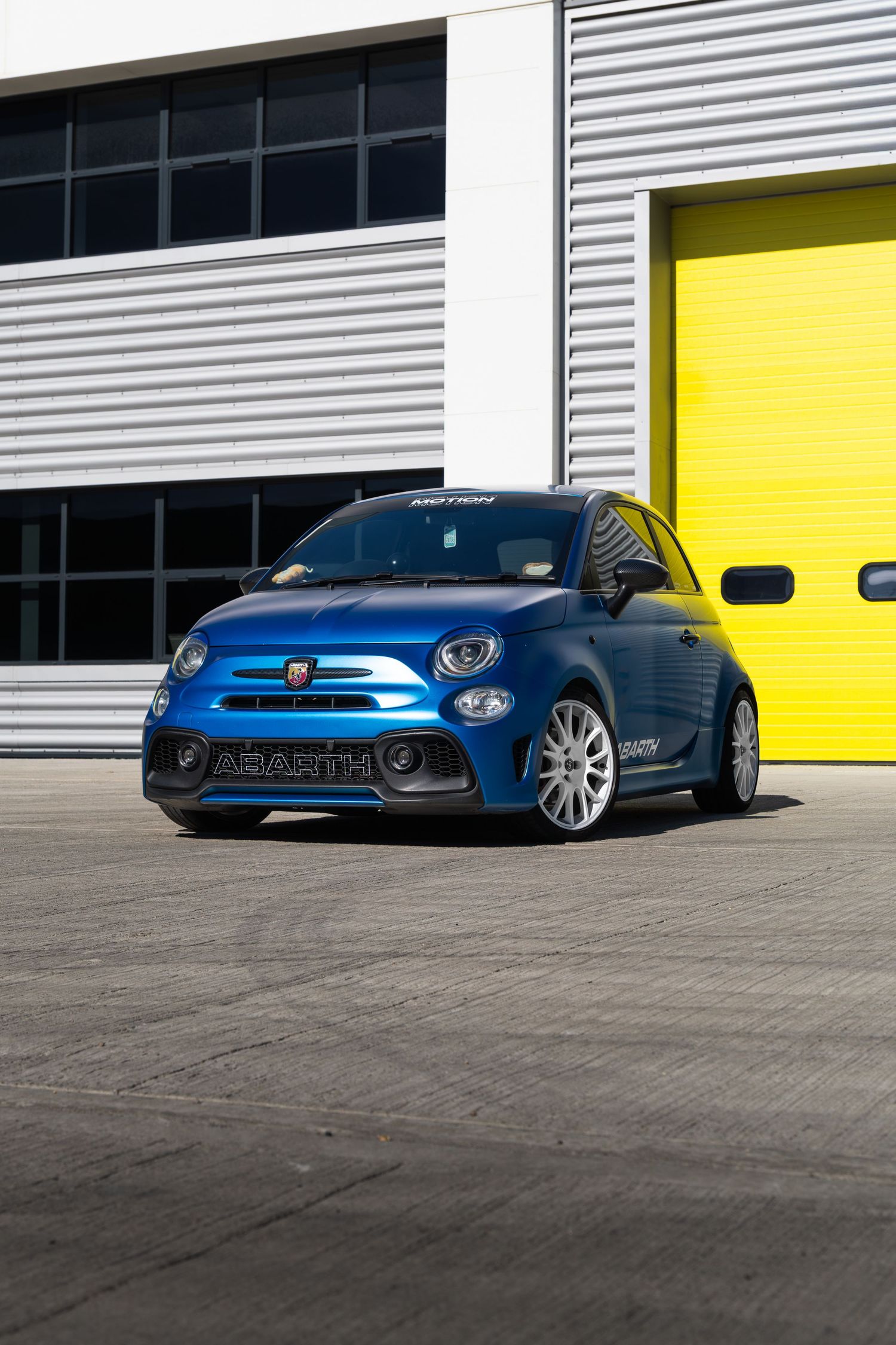 Blue Abarth 500 hatchback posed in front of bright yellow roller doors at a racing circuit.