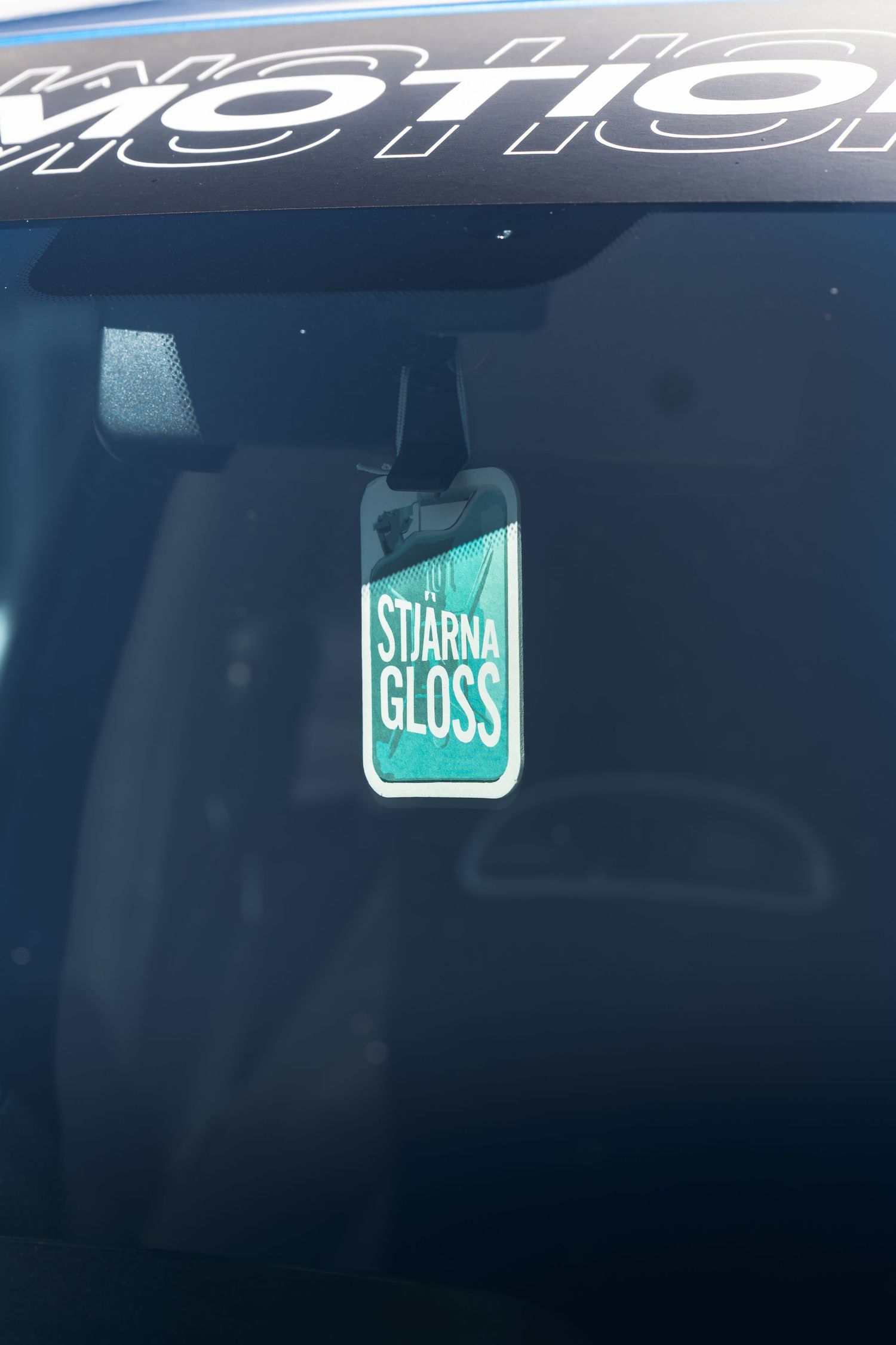 Stjarna Gloss air freshener hanging from rearview mirror inside a dark car interior.