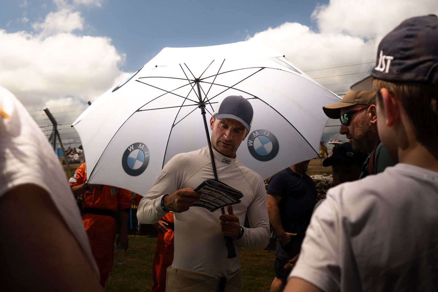 A racing driver in white firesuit signs autographs under a BMW umbrella at an event.