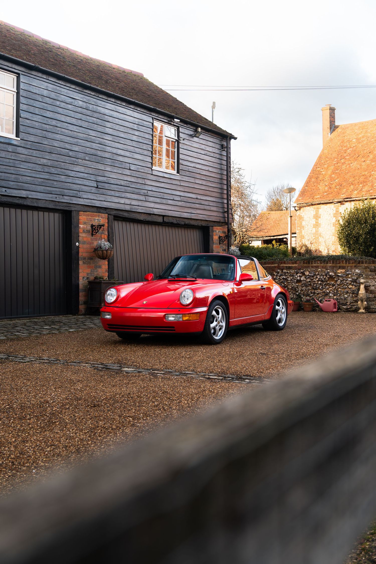 Red Porsche 911 Carrera 2 Targa parked on gravel driveway beside a dark timber-clad garage building.