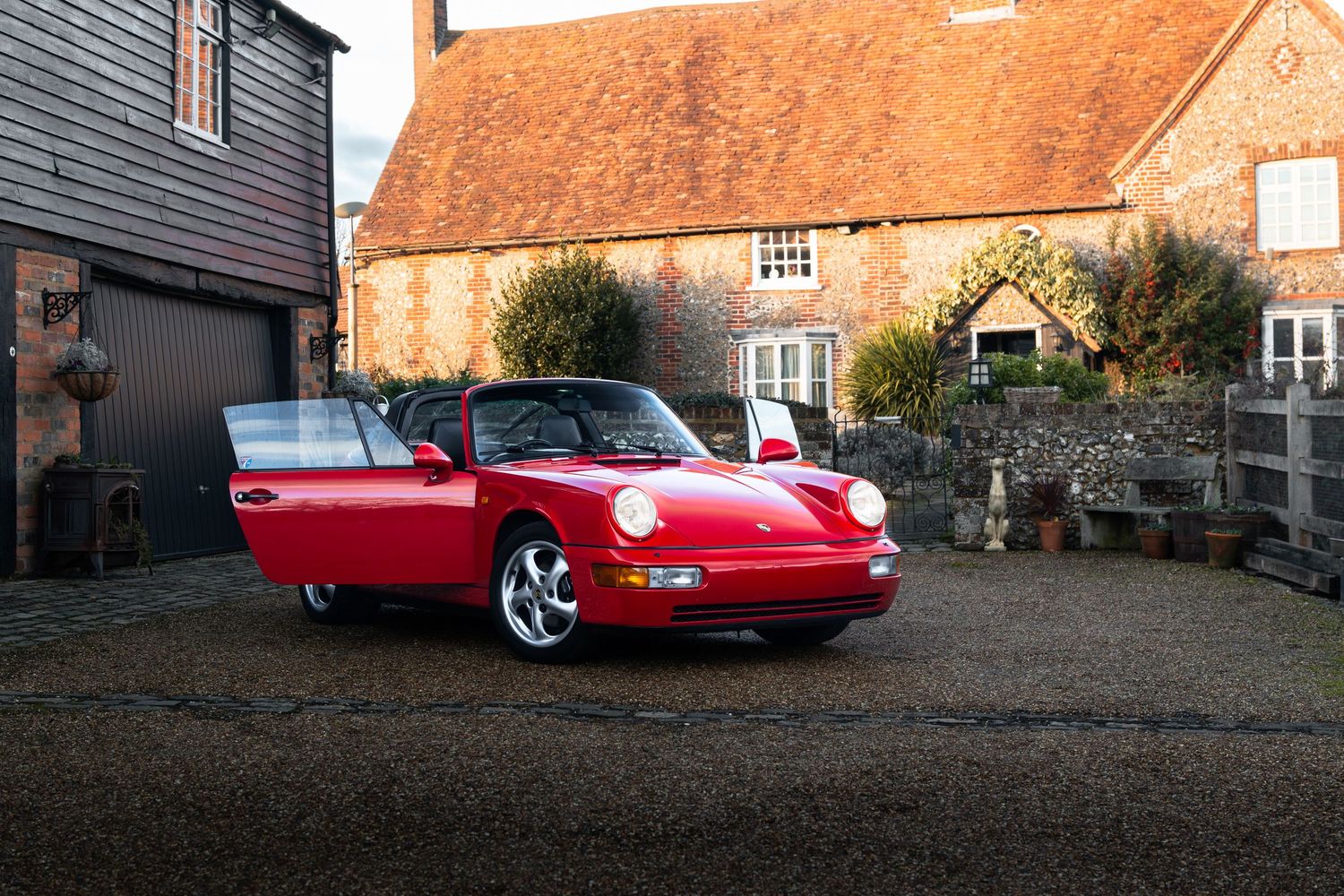 Red Porsche 911 Carrera convertible parked on gravel driveway beside rustic brick farmhouse building.
