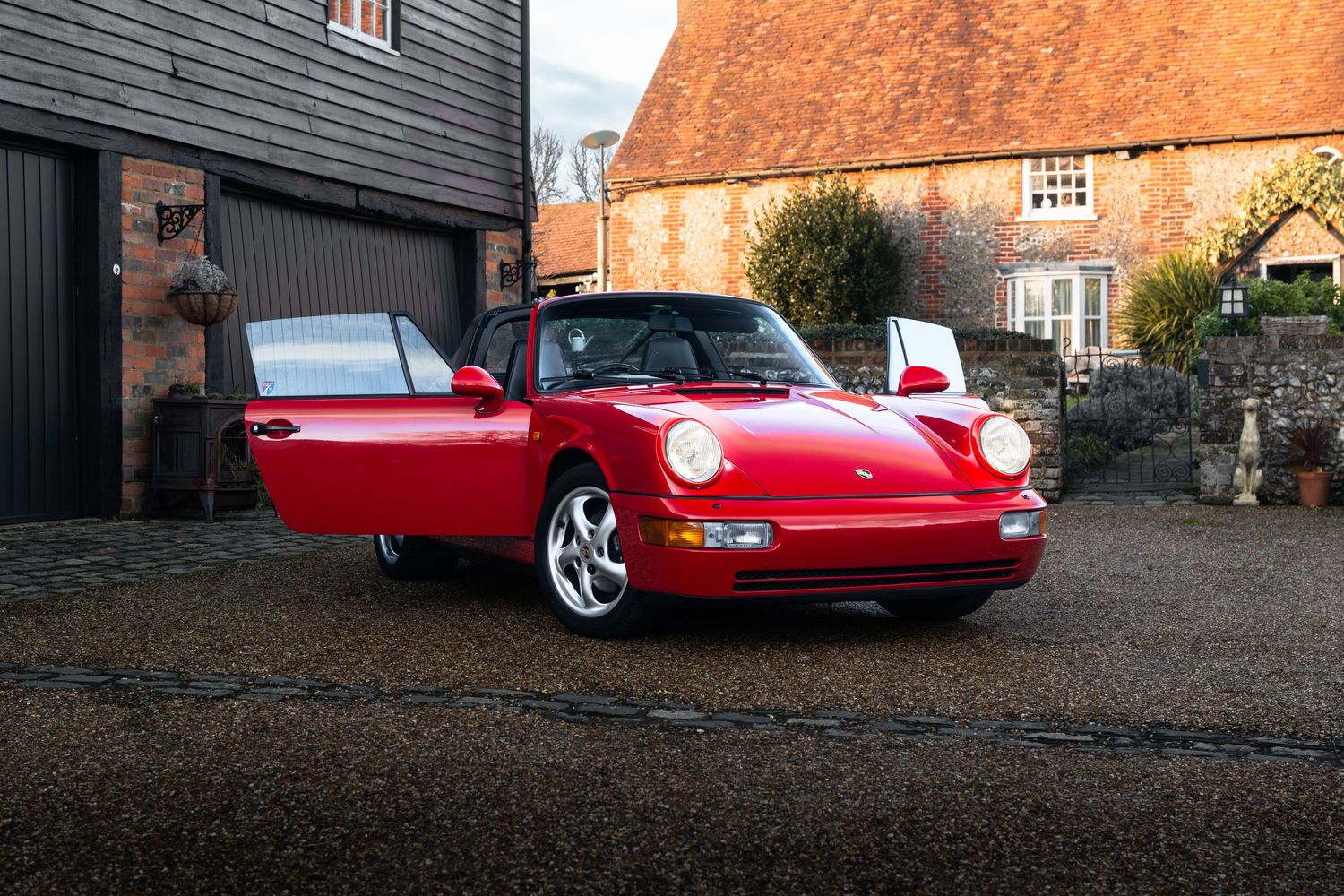 Guards Red Porsche 911 964 Carrera Targa with open roof parked on gravel outside a rustic English farmhouse.