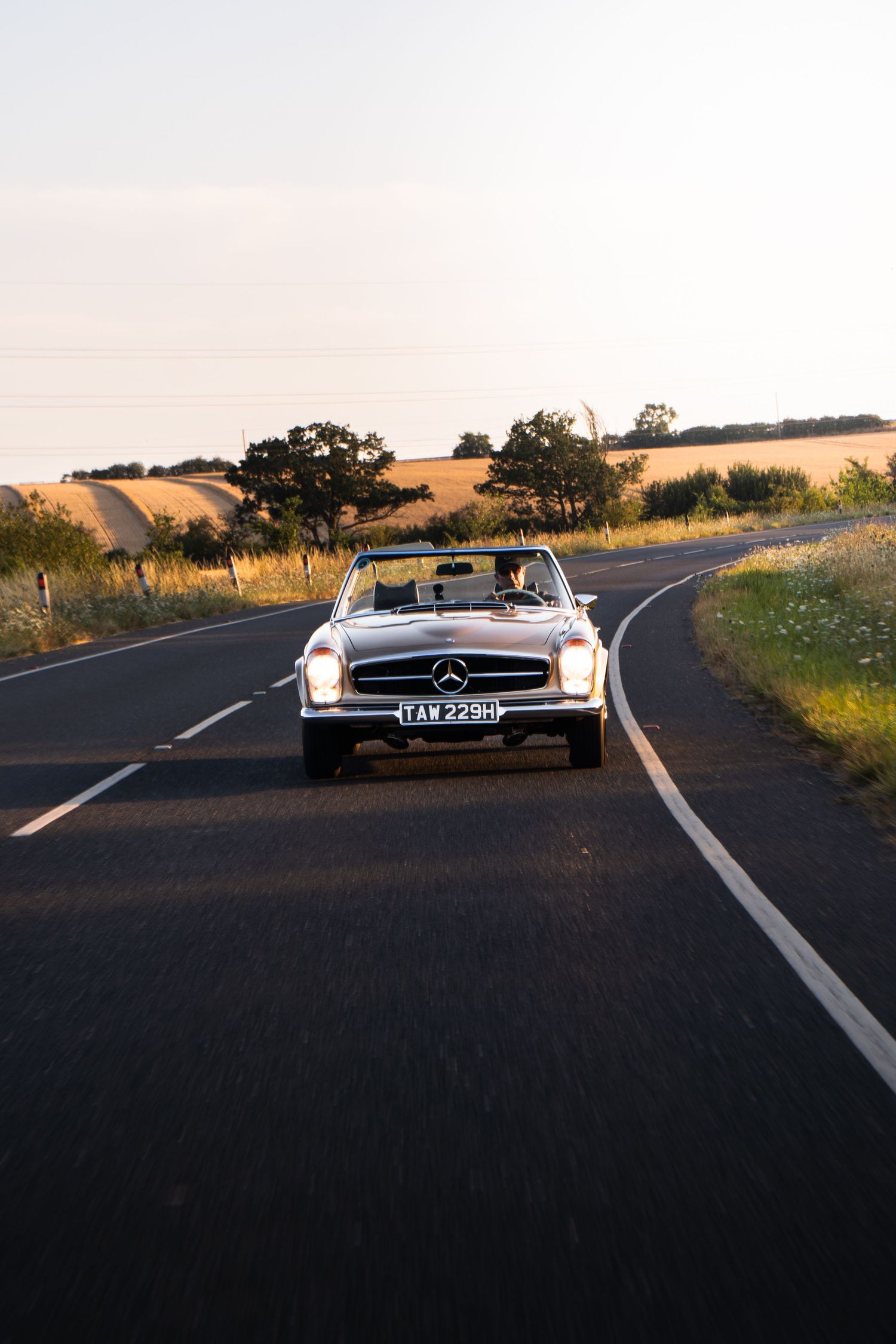 Classic Mercedes-Benz convertible driving on a scenic rural road at golden hour, surrounded by fields and trees.