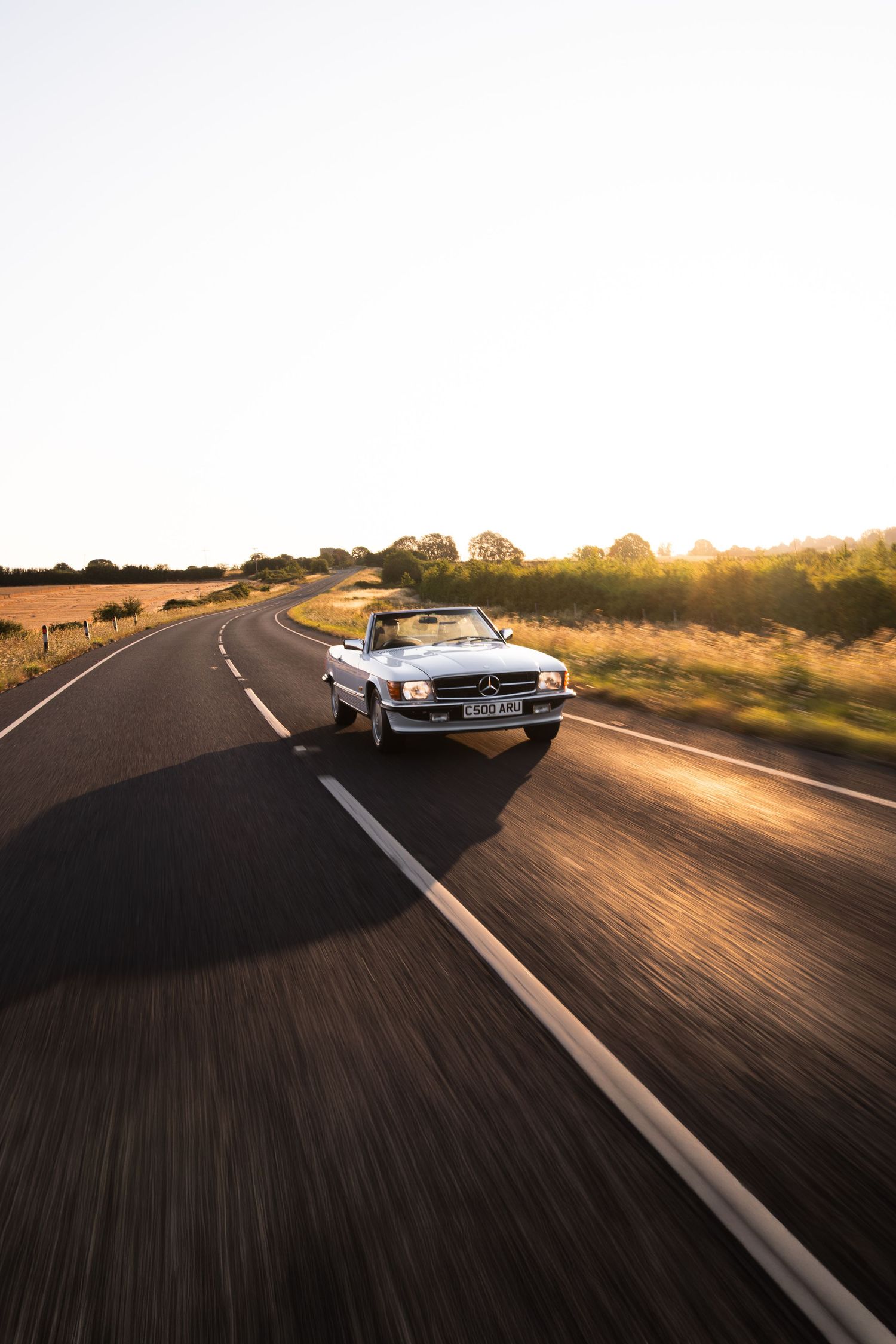 White classic Mercedes-Benz convertible driving on a rural road at golden hour sunset.