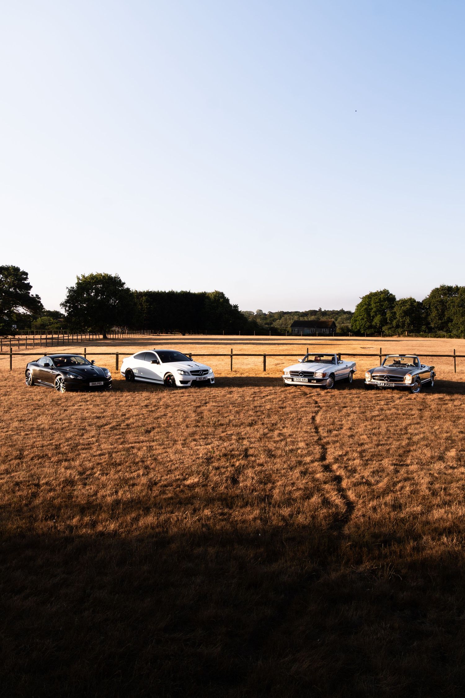 Four luxury cars parked in a golden harvested field at sunset with trees and blue sky in the background.