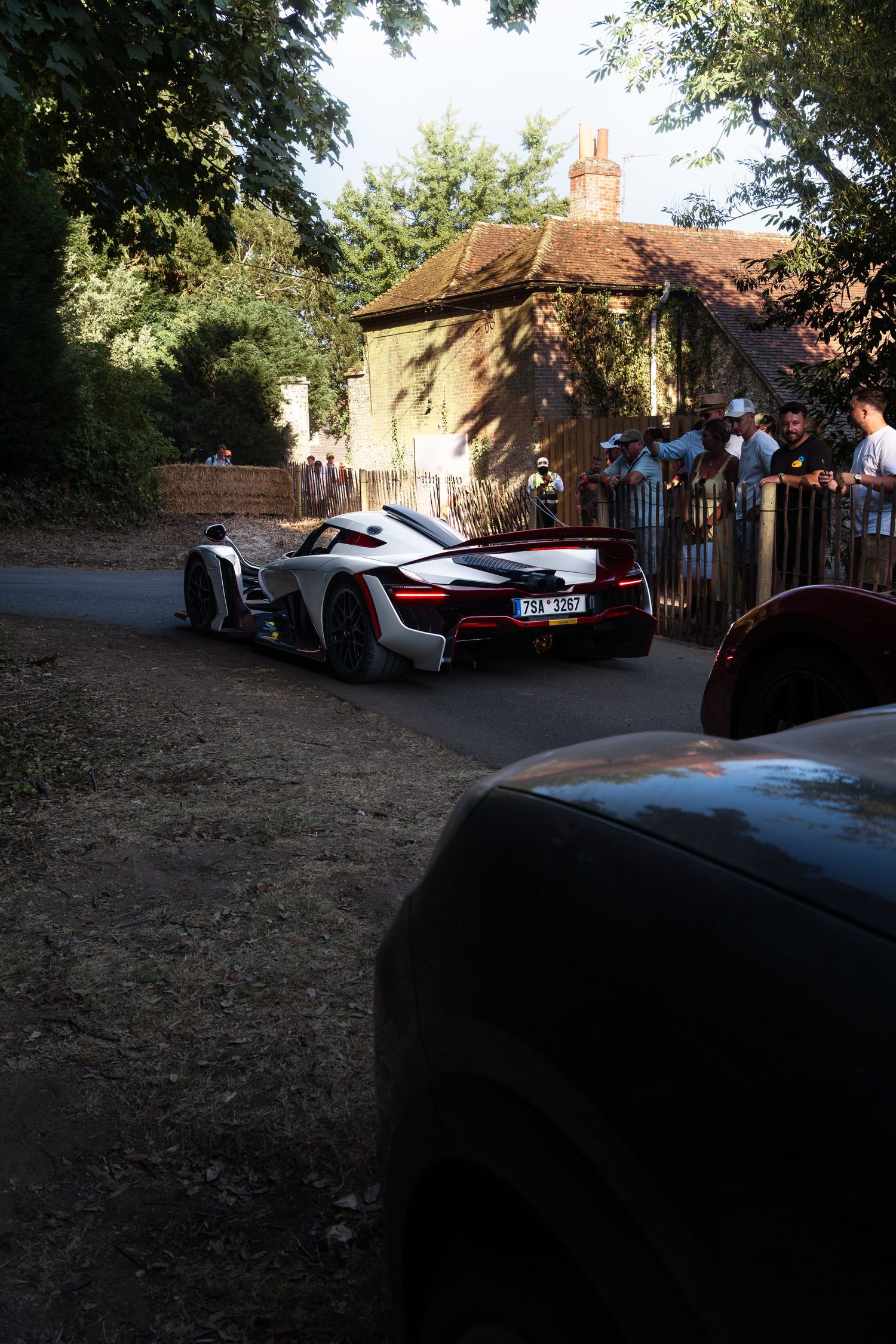 A sleek supercar drives past spectators at the Goodwood Festival of Speed, surrounded by trees and a historic building.