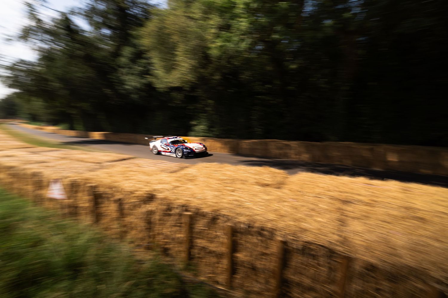 Red Bull racing car speeds past hay bales on a tree-lined hillclimb course at Goodwood Festival of Speed.