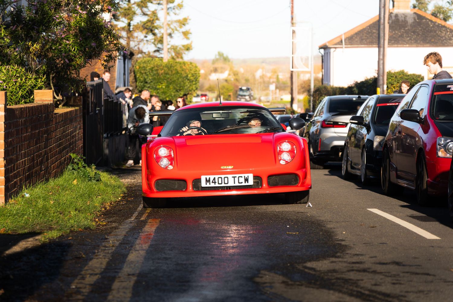 Red Noble supercar with UK license plate driving through a residential street.
