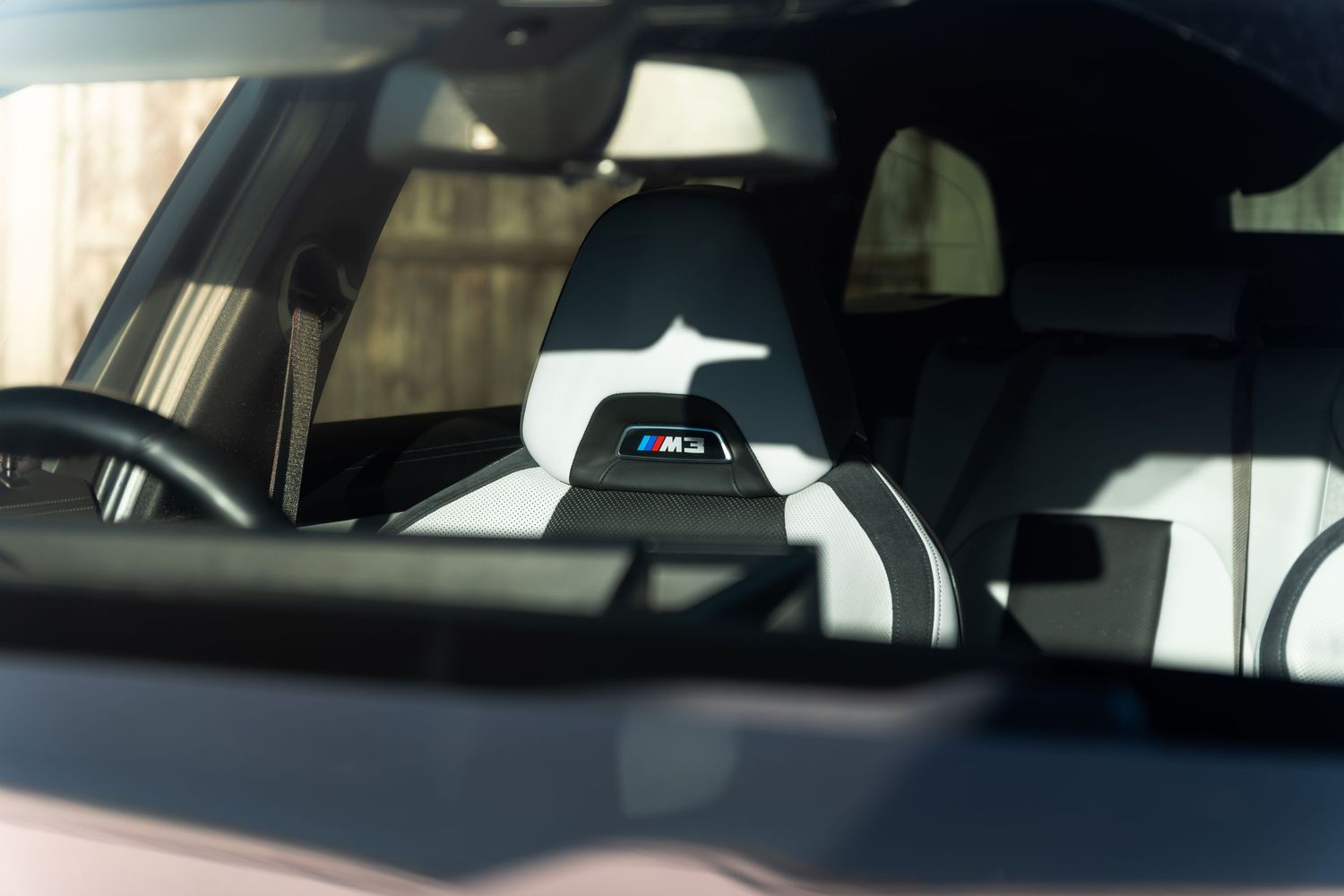 Interior view through windshield of a BMW M3 showing M3 branded headrest and sunlit leather seats.
