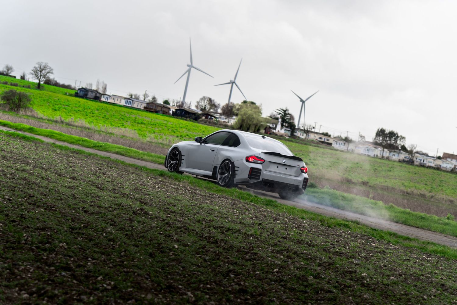Silver BMW M2 coupe performing a smoky burnout on a rural road with wind turbines in the background.