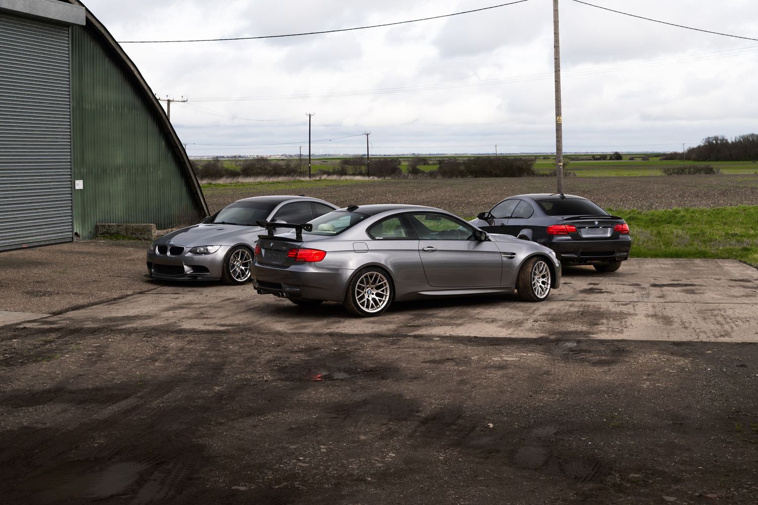Three BMW M3 E92 coupes parked near a large green metal barn on a rural gravel lot.