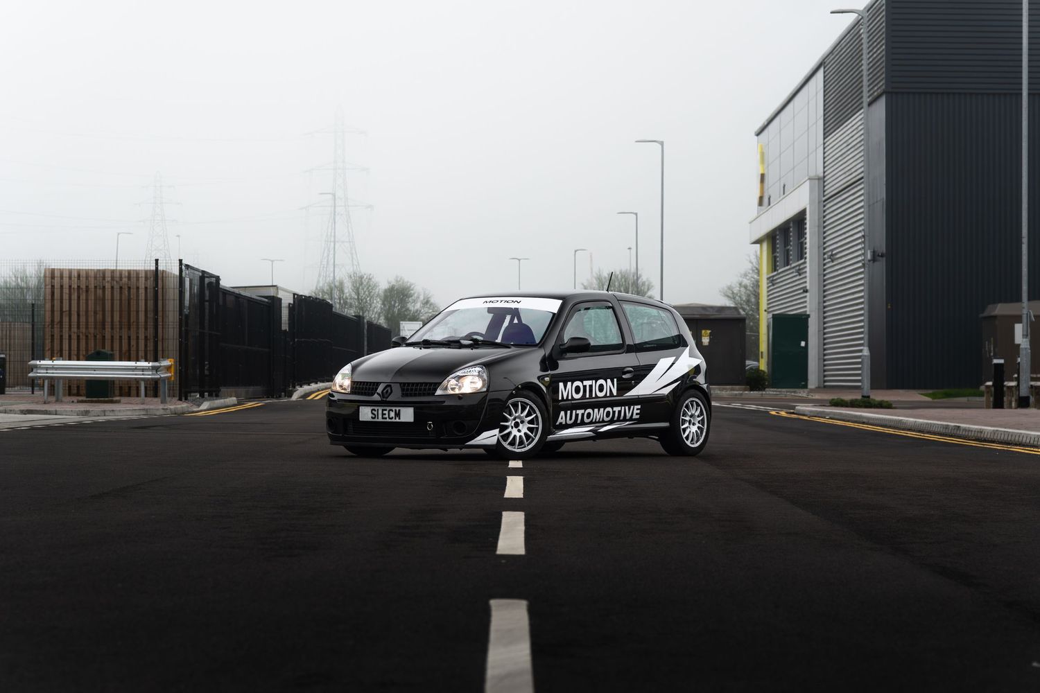 Black Motion Automotive liveried Renault Clio centered on a road in a foggy industrial estate setting.