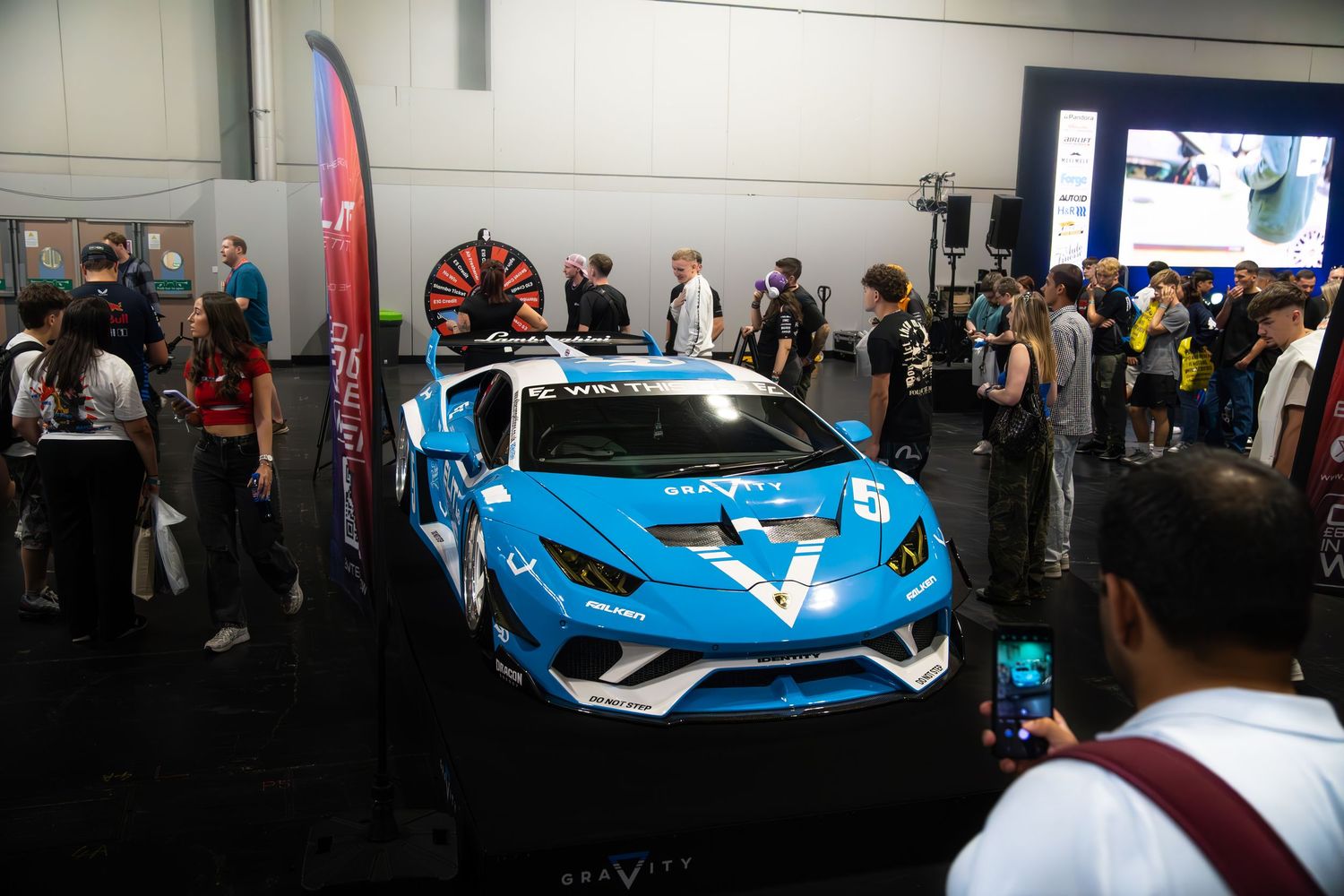 Blue Gravity-sponsored Lamborghini race car surrounded by crowds at an indoor automotive expo.