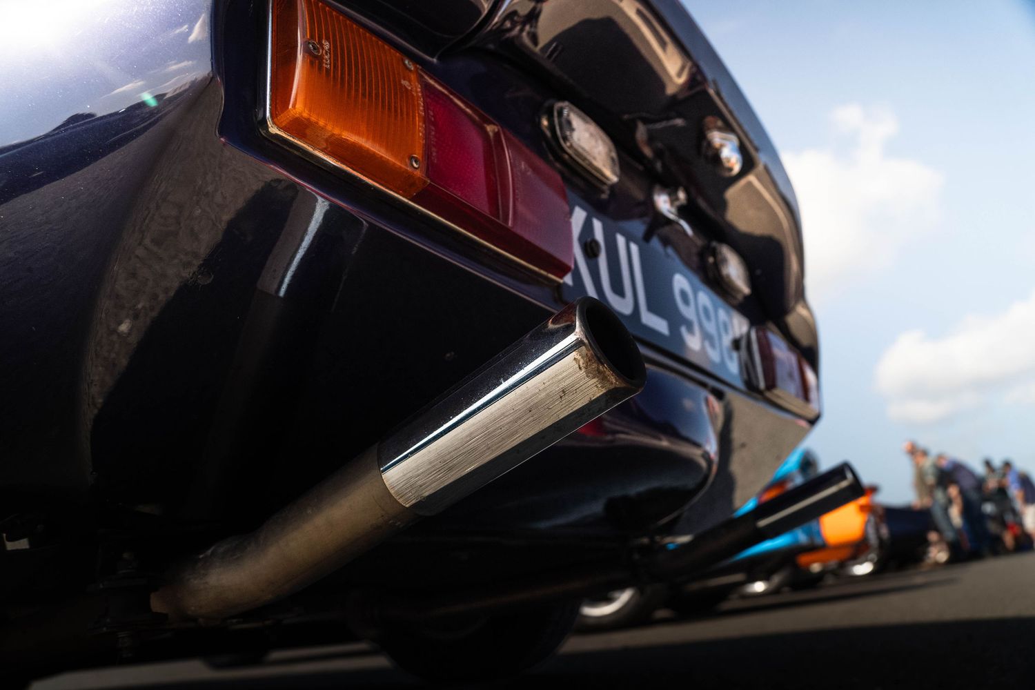 Close-up of rear bumper and exhaust of a classic car with UL 998 number plate at a race event.