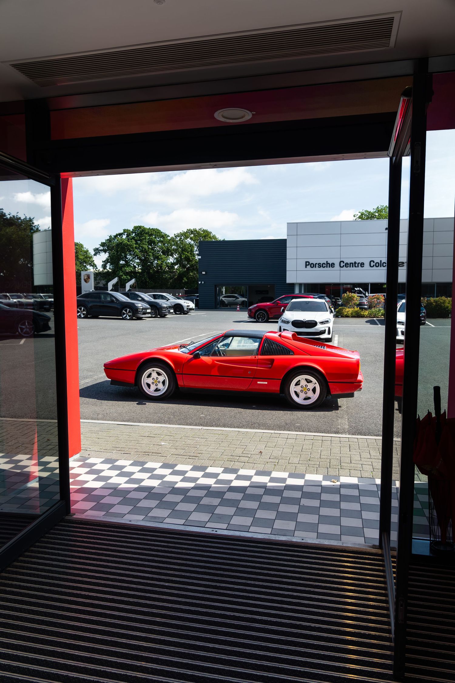 Red Ferrari 328 GTS sports car parked outside a Porsche Centre dealership on a checkered floor entrance.
