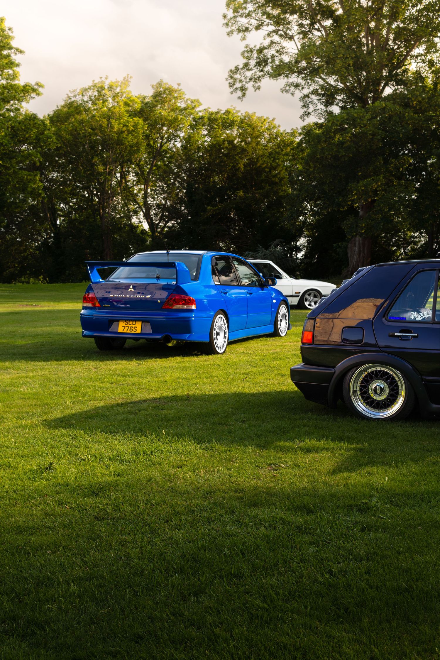 Blue Mitsubishi Lancer Evolution parked on green grass at a car show with other modified cars and trees in background.