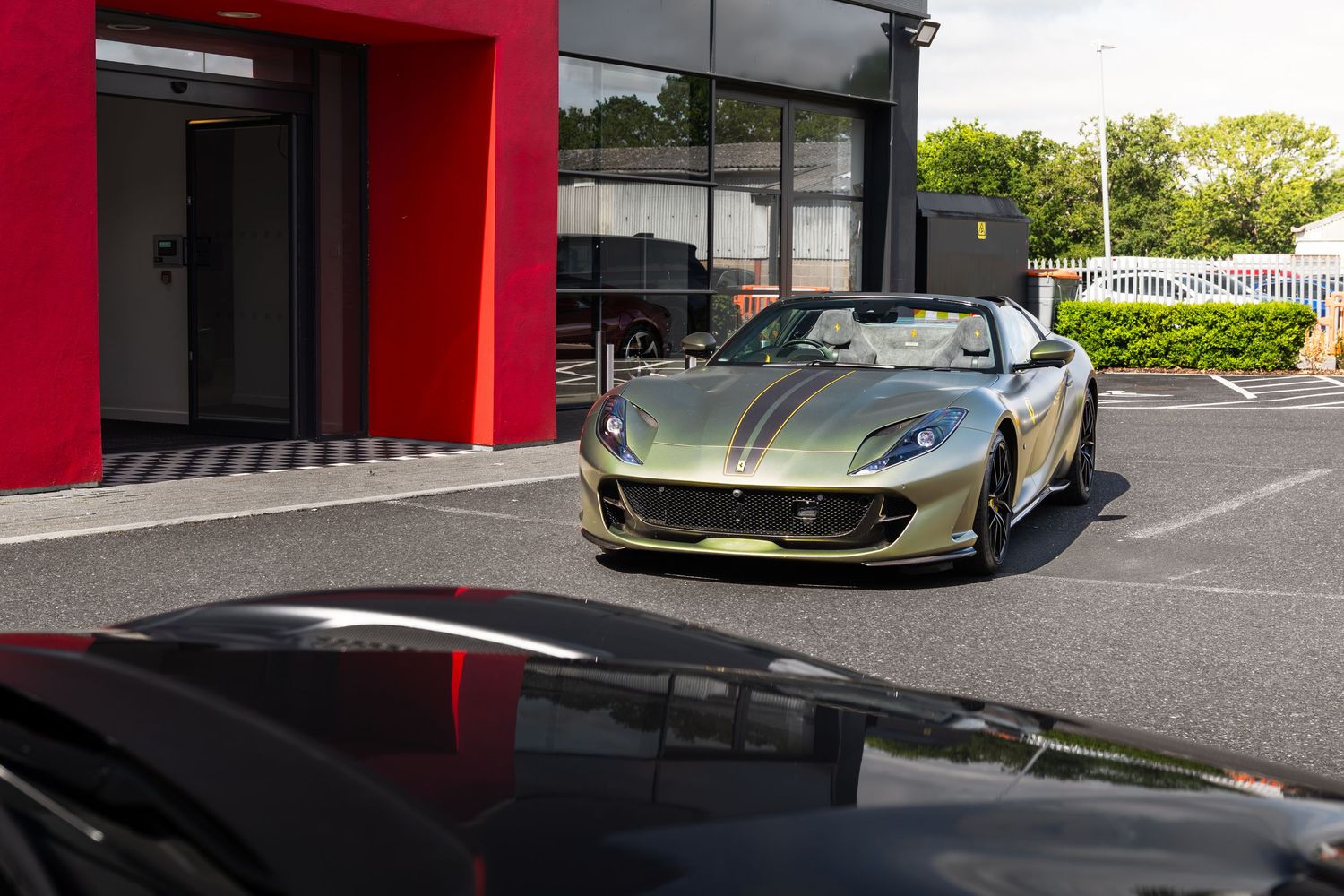 Gold Ferrari 812 GTS spider parked outside a Ferrari dealership on a sunny day.