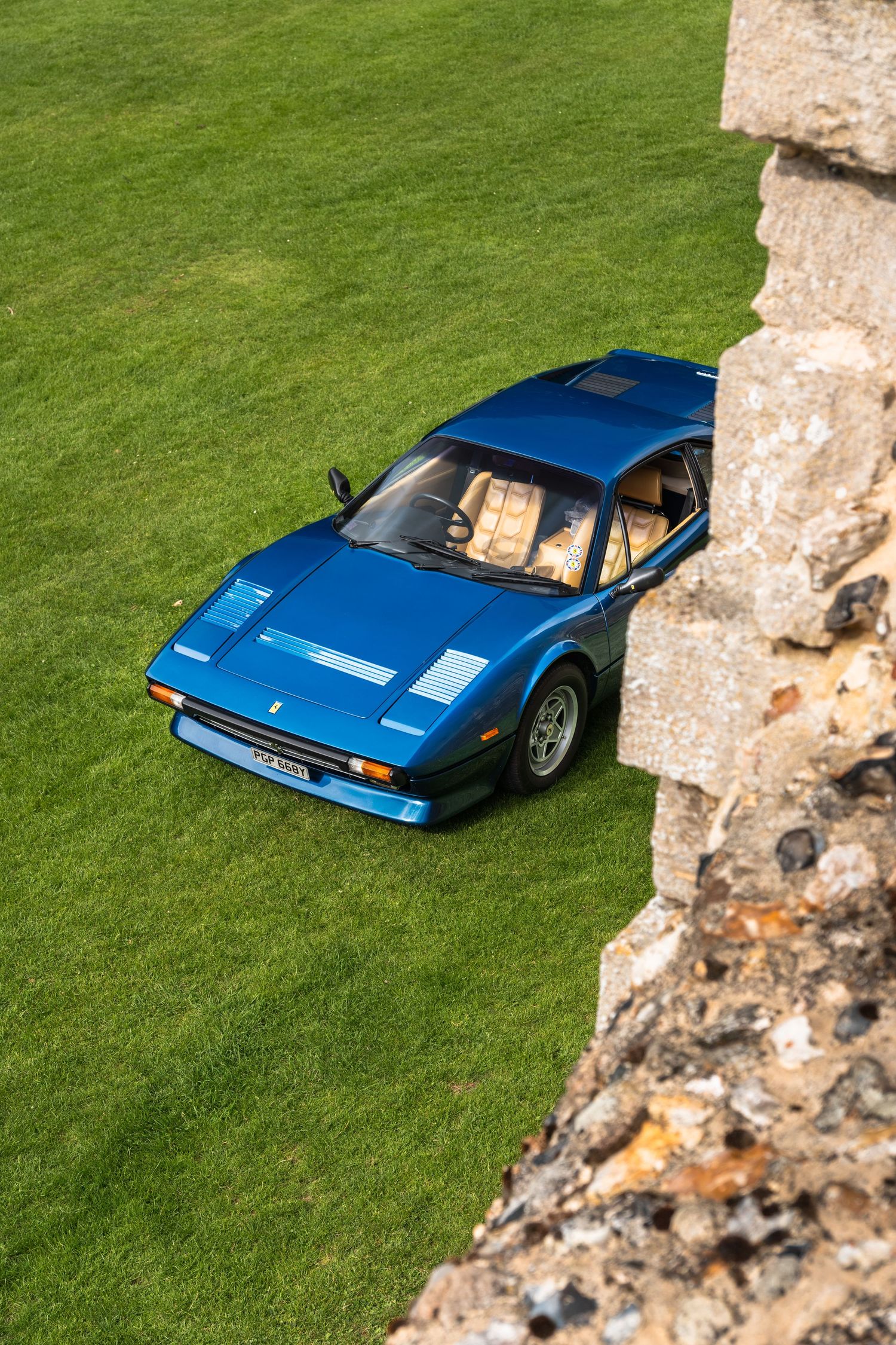 Blue Ferrari 308 GT4 sports car parked on green grass beside a stone wall, photographed from above.