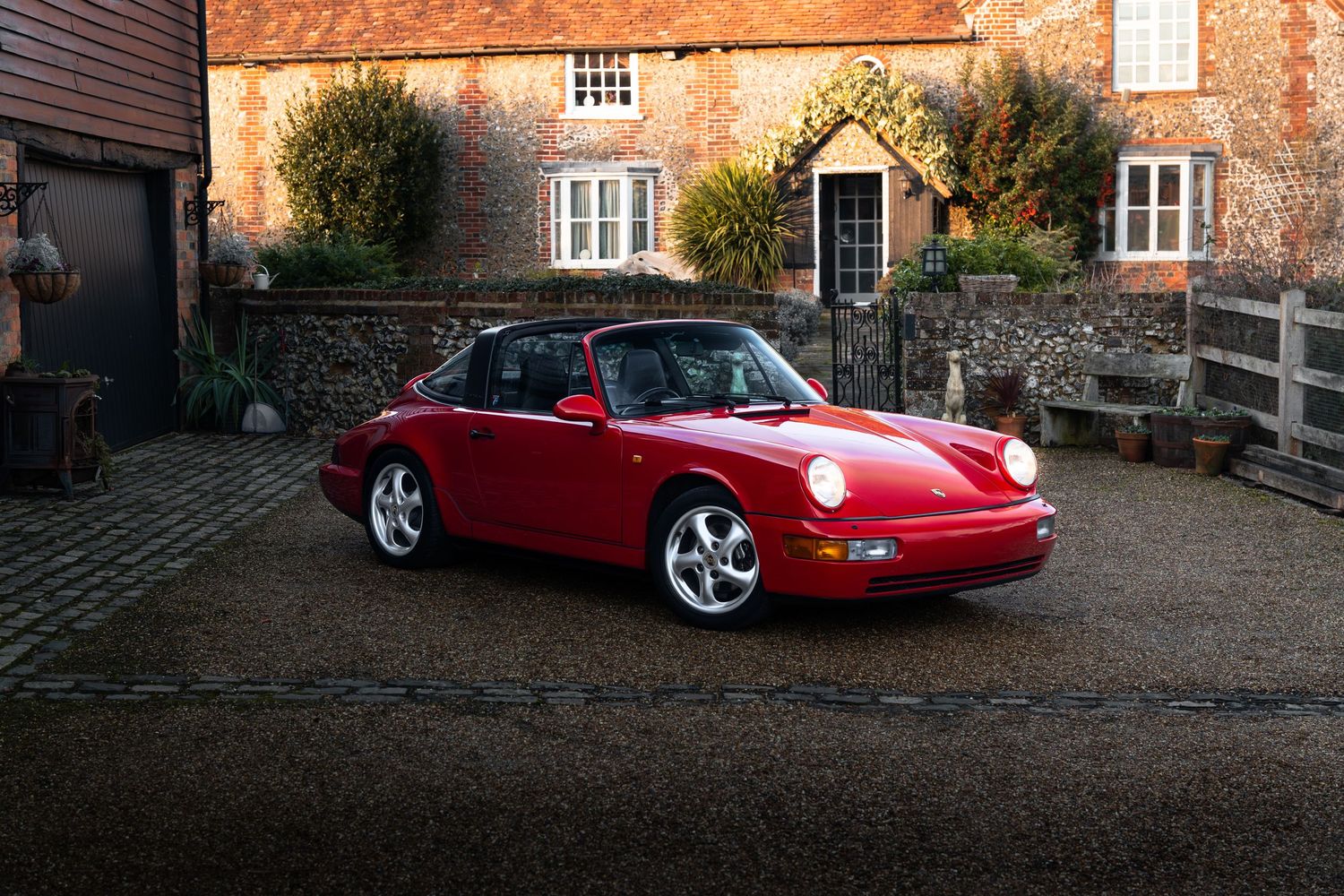 Vintage red Porsche 911 Targa parked on cobblestone driveway beside charming English countryside building.